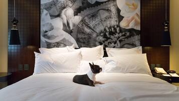 Una habitación de hotel con una cama, ropa de cama blanca, un perro, un tapiz floral grande en la pared y una mesita de noche con un teléfono y una lámpara.