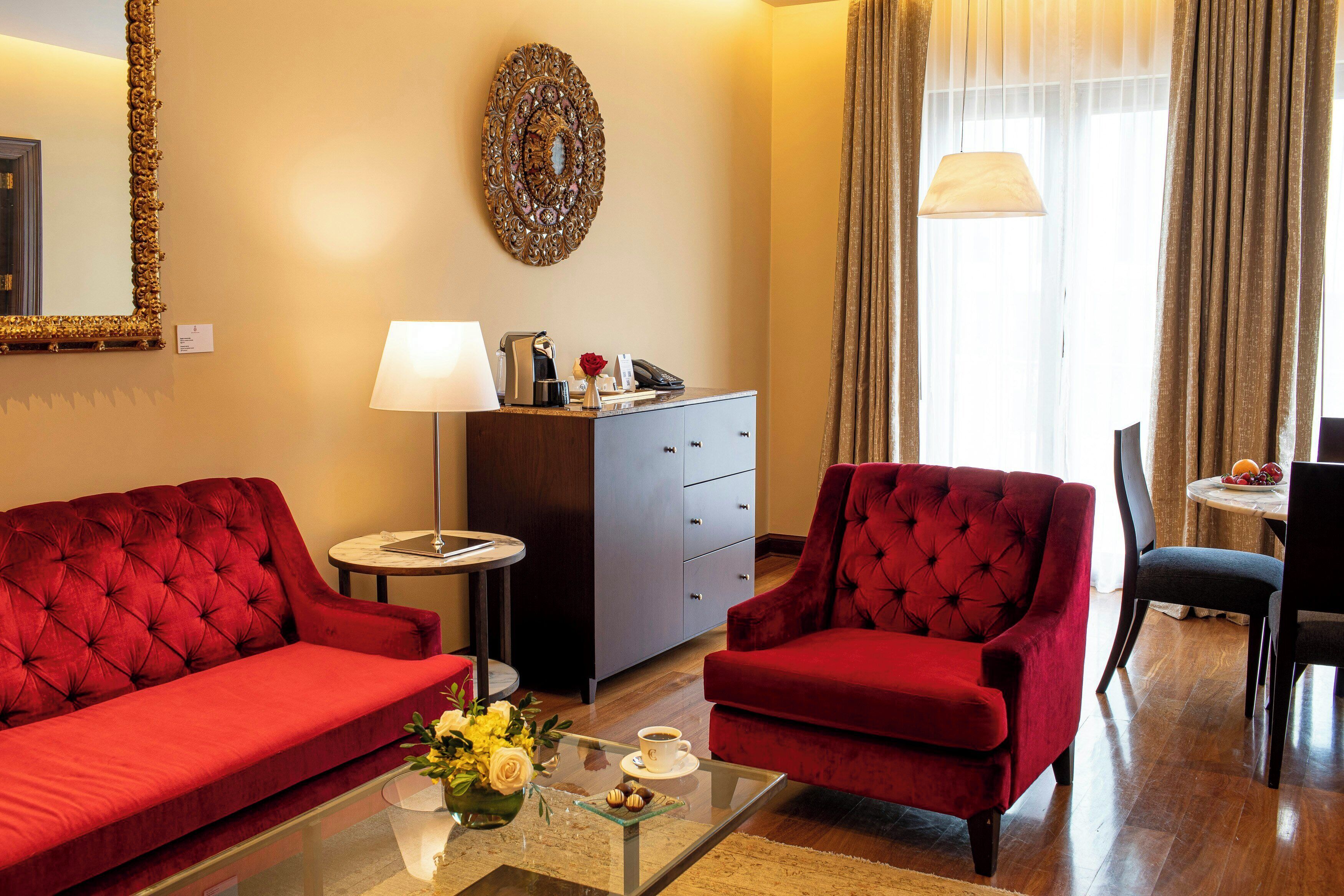 A hotel room with a red sofa, a red armchair, a small table with a vase of flowers, a lamp, and a mirror on the wall.