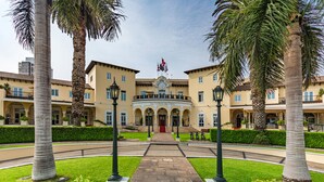Entrada de hotel con un camino central, palmeras y un edificio de múltiples ventanales y una bandera.