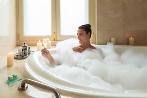 Eine Person liegt in einer Badewanne voller Blasen, mit Kerzen und Pflegeprodukten daneben.