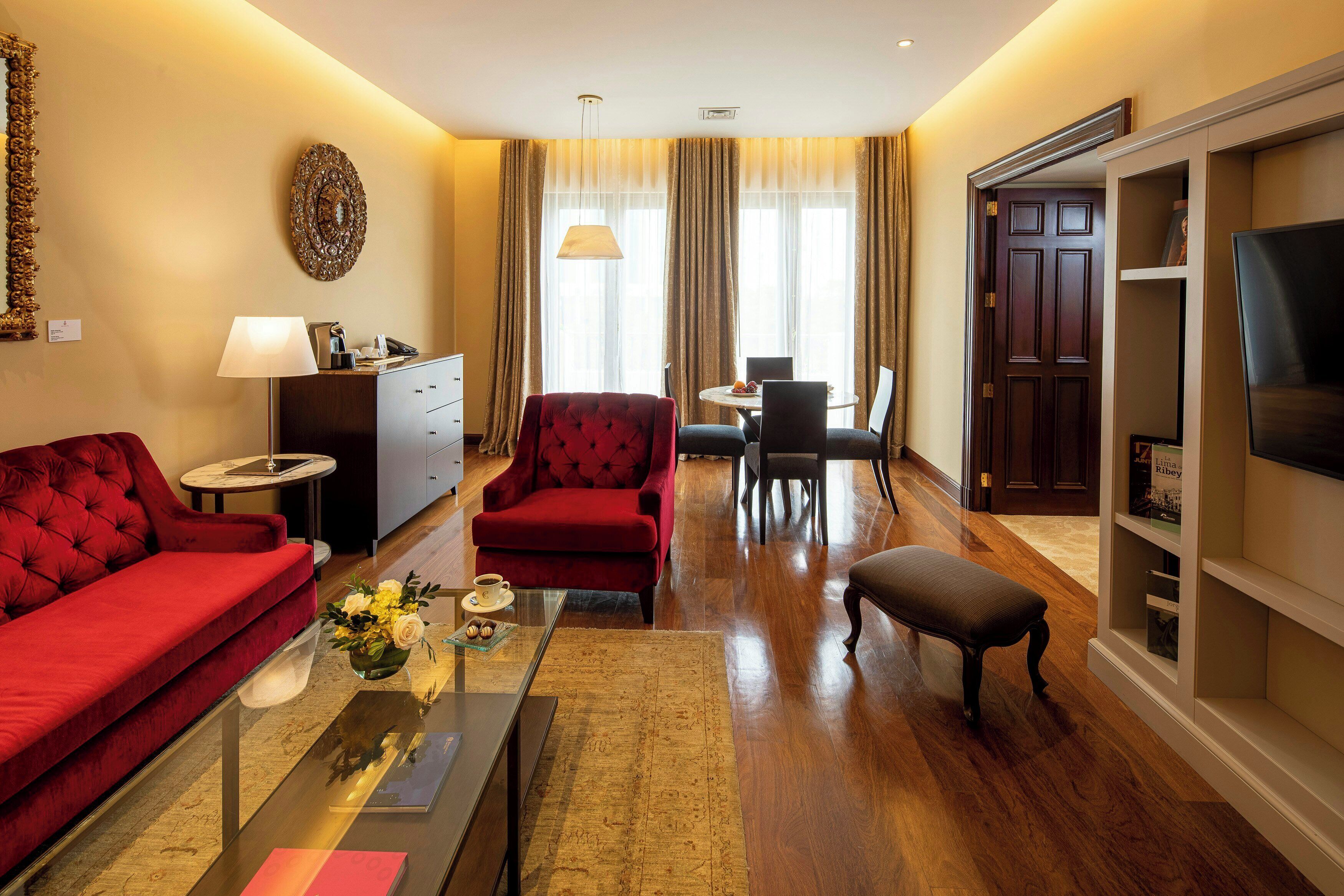 Habitación de hotel con sofá rojo, sillón rojo, mesa de comedor con sillas, televisor y estantería.