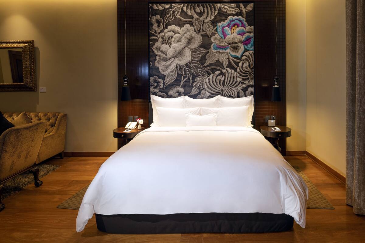Ein großes Bett mit weißer Bettwäsche, ein dunkles Kopfteil mit floralen Motiven, zwei Nachttische mit Lampen und ein gepolsterter Ohrensessel.