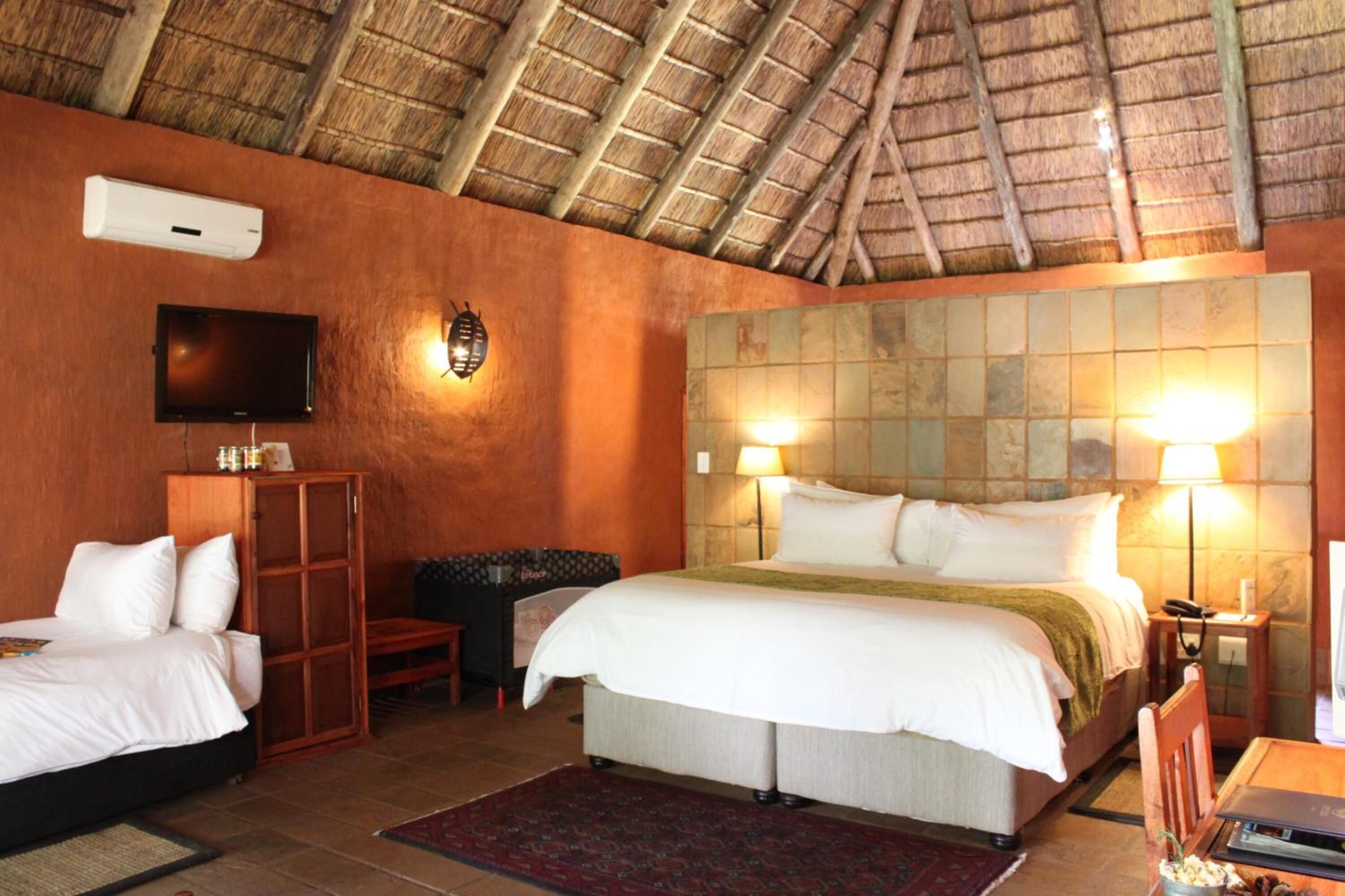 A hotel room with a thatched roof, a large bed with white linens, a television mounted on the wall, and a wooden cabinet.