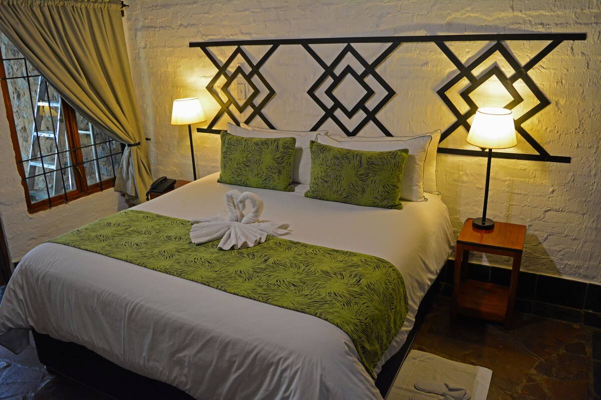 A neatly made bed with a green and white bedspread, two green pillows, and a white towel on the bed. There are two bedside lamps on wooden nightstands.