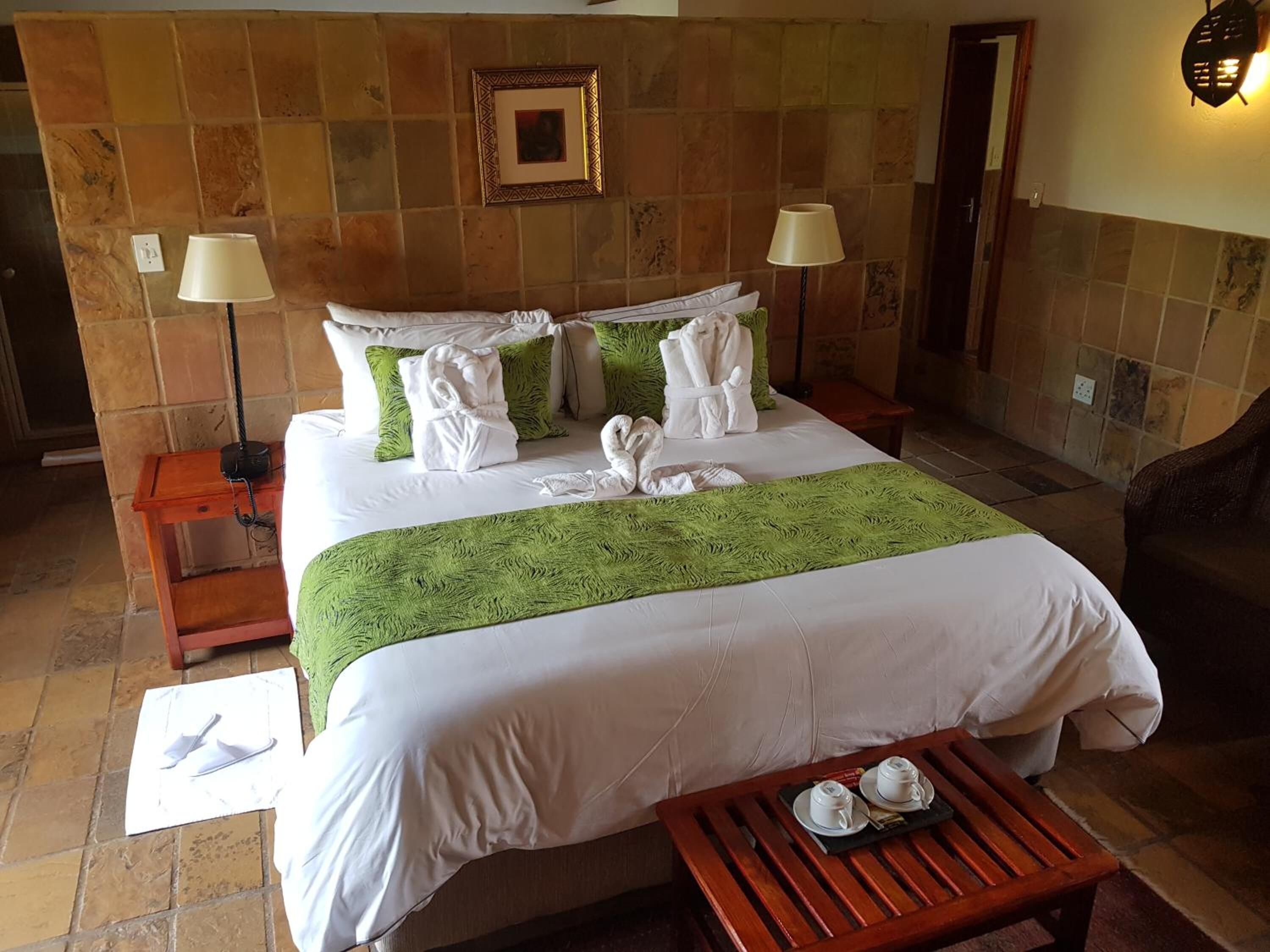 A neatly made bed with white linens, green throw, and towels, a wooden nightstand with a lamp, and a small table with cups and saucers.