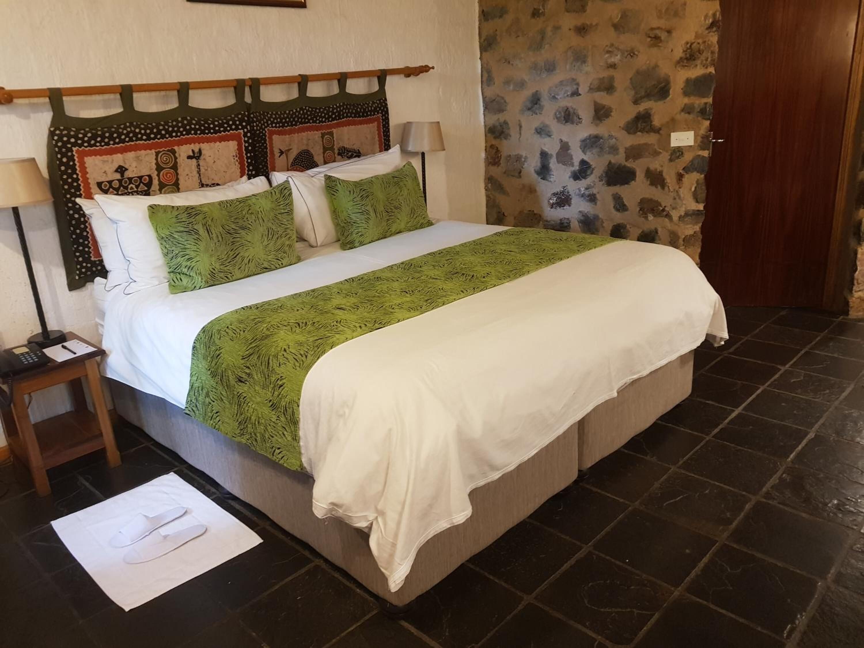 A neatly made bed with a green and white bedspread, a wooden headboard with decorative panels, a bedside table with a lamp, and a stone wall.