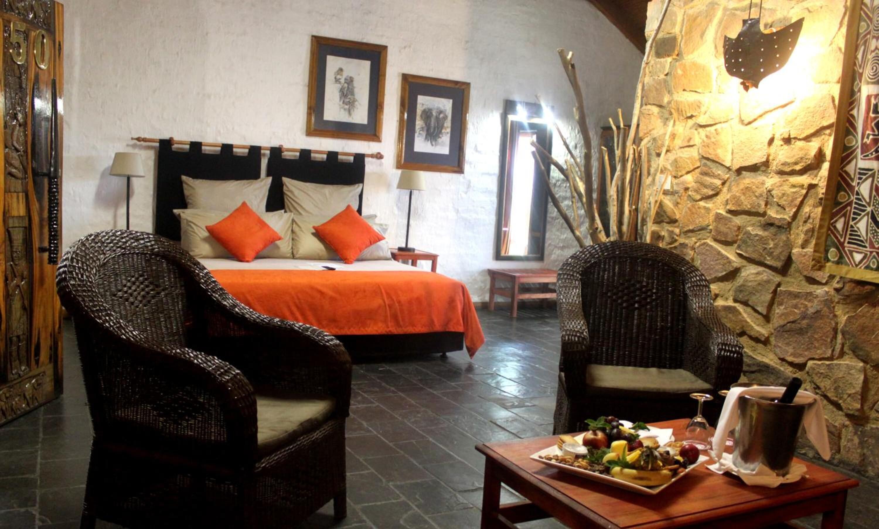 A bedroom with a bed, two chairs, a table with fruit and a drink, and a stone wall.