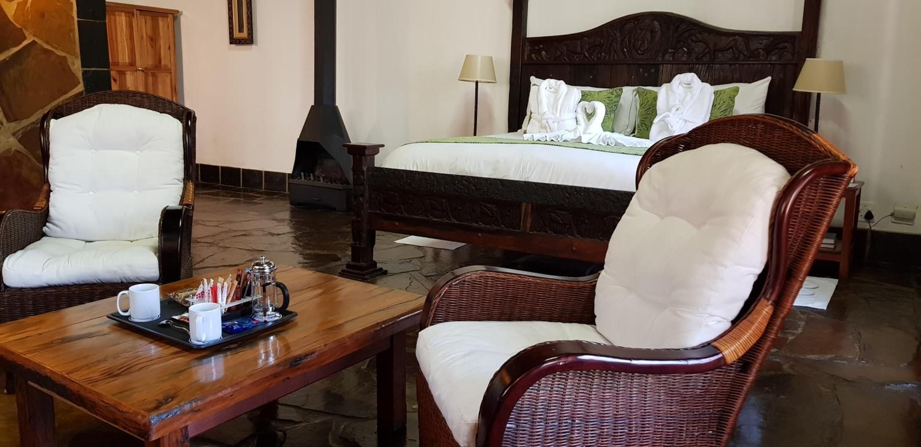 A hotel room with a bed, two wicker chairs, a wooden table with a tray of items, and a fireplace.