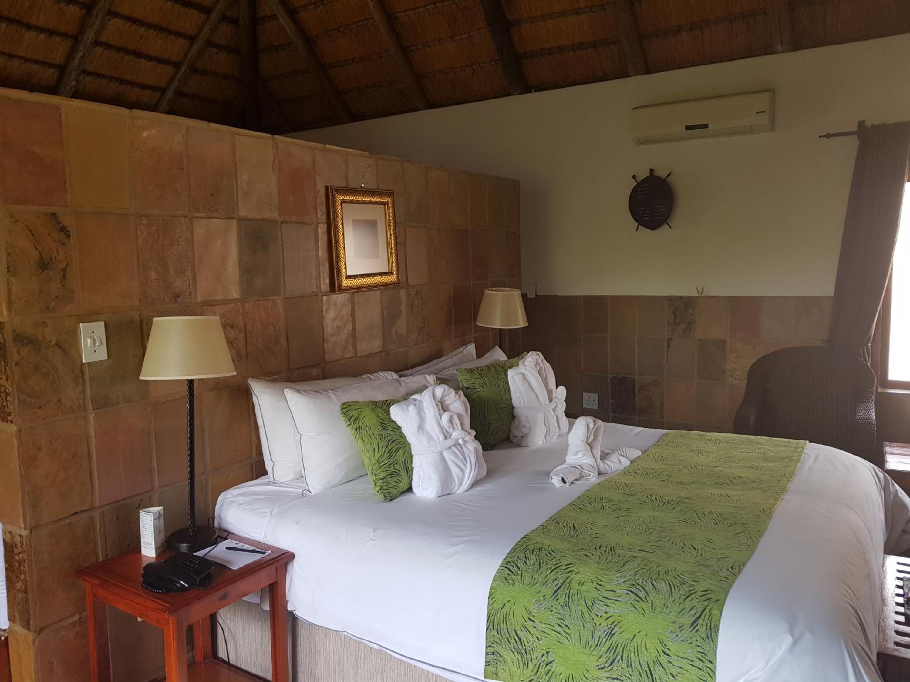 A hotel room with a stone wall, a bed with white bedding and green throw pillows, two bedside lamps, a small wooden nightstand with a phone and papers, and a wall-mounted air conditioning unit.