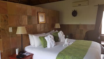 A hotel room with a stone wall, a bed with white bedding and green throw pillows, two bedside lamps, a small wooden nightstand with a phone and papers, and a wall-mounted air conditioning unit.