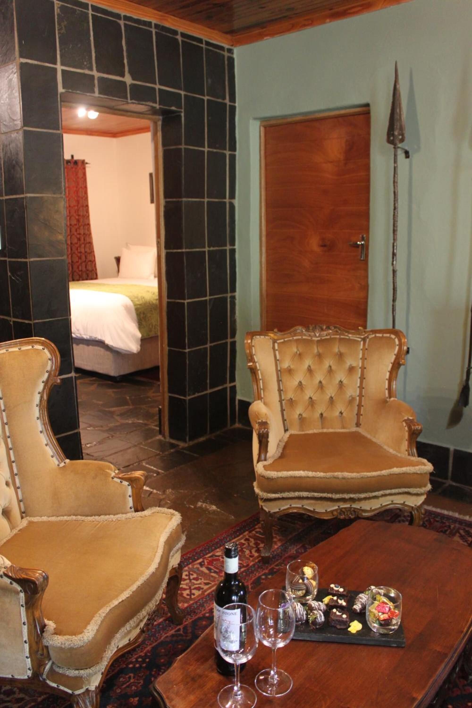 A room with two plush armchairs, a wooden table with a bottle and glasses, and a bed in the background.