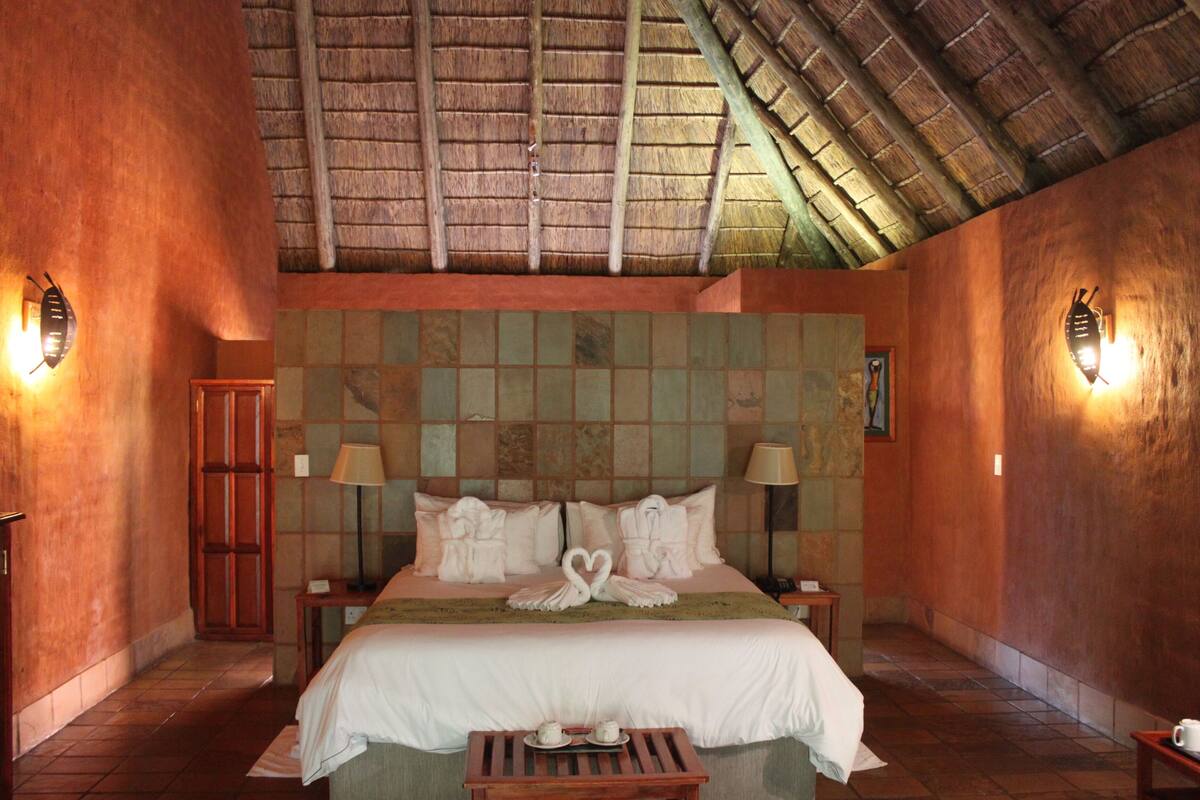 A hotel room with a large bed, two bedside tables, a nightstand, a wooden bench, and a thatched roof ceiling.