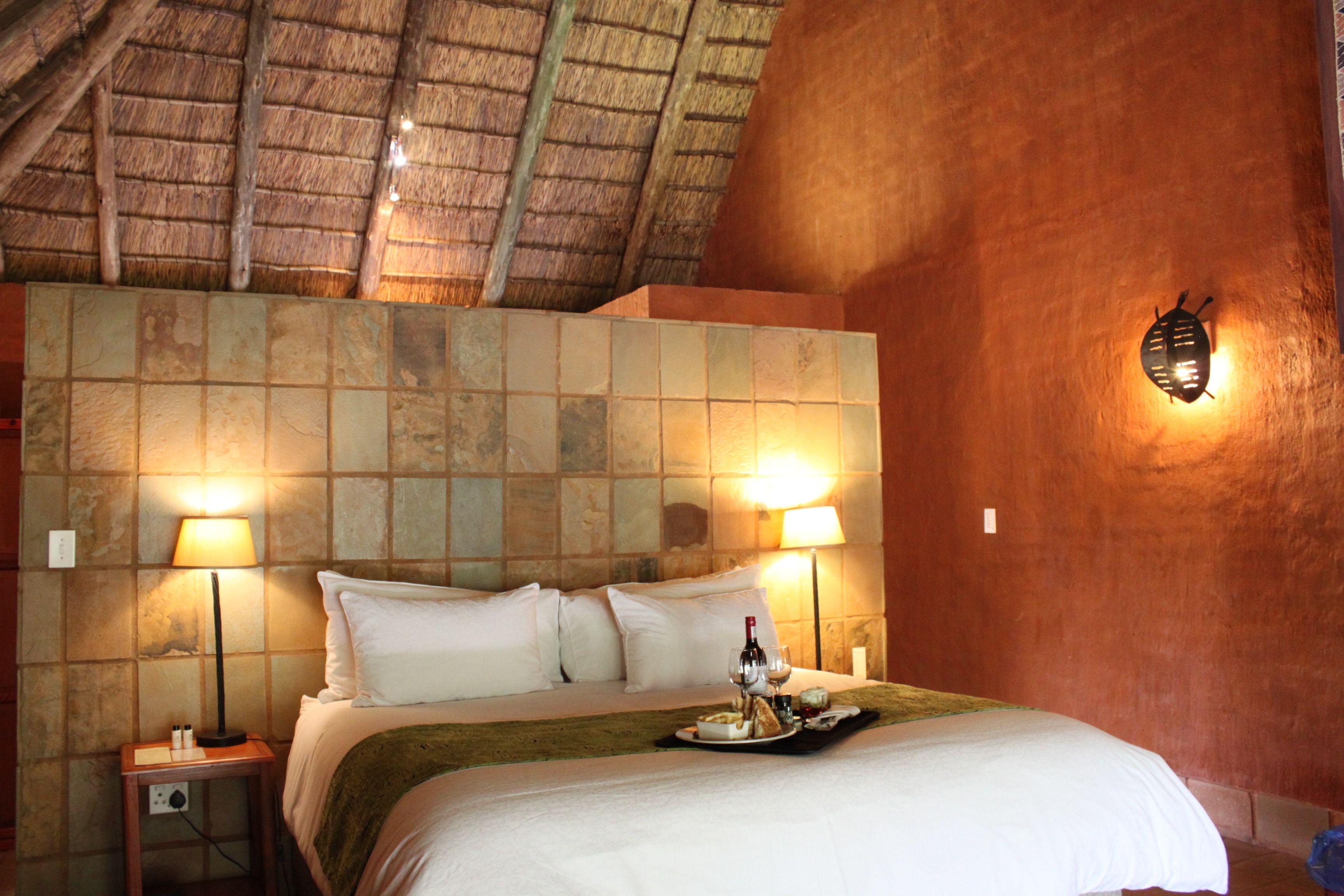 Une chambre d’hôtel avec un grand lit, deux lampes de chevet, un plateau avec des verres à vin et une bouteille, et un mur en pierre à la texture particulière.