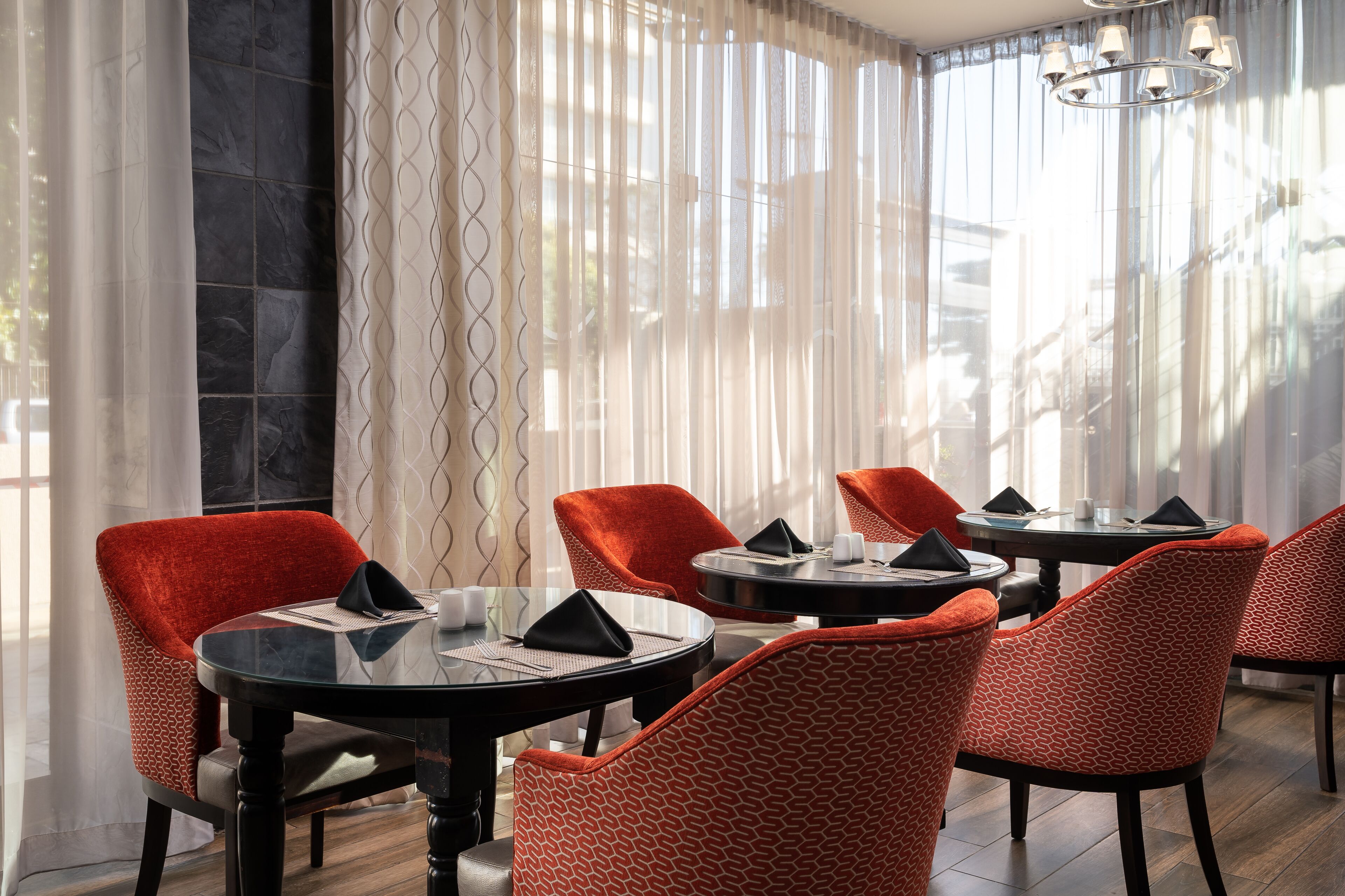 A dining area with round tables and orange chairs, large windows with sheer curtains, and a chandelier.