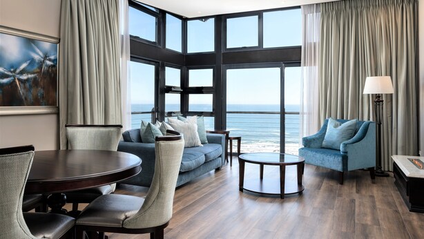 A modern living room with a large window overlooking the sea, a round dining table, and comfortable seating. - Premier Hotel Cape Town (Cape Town)