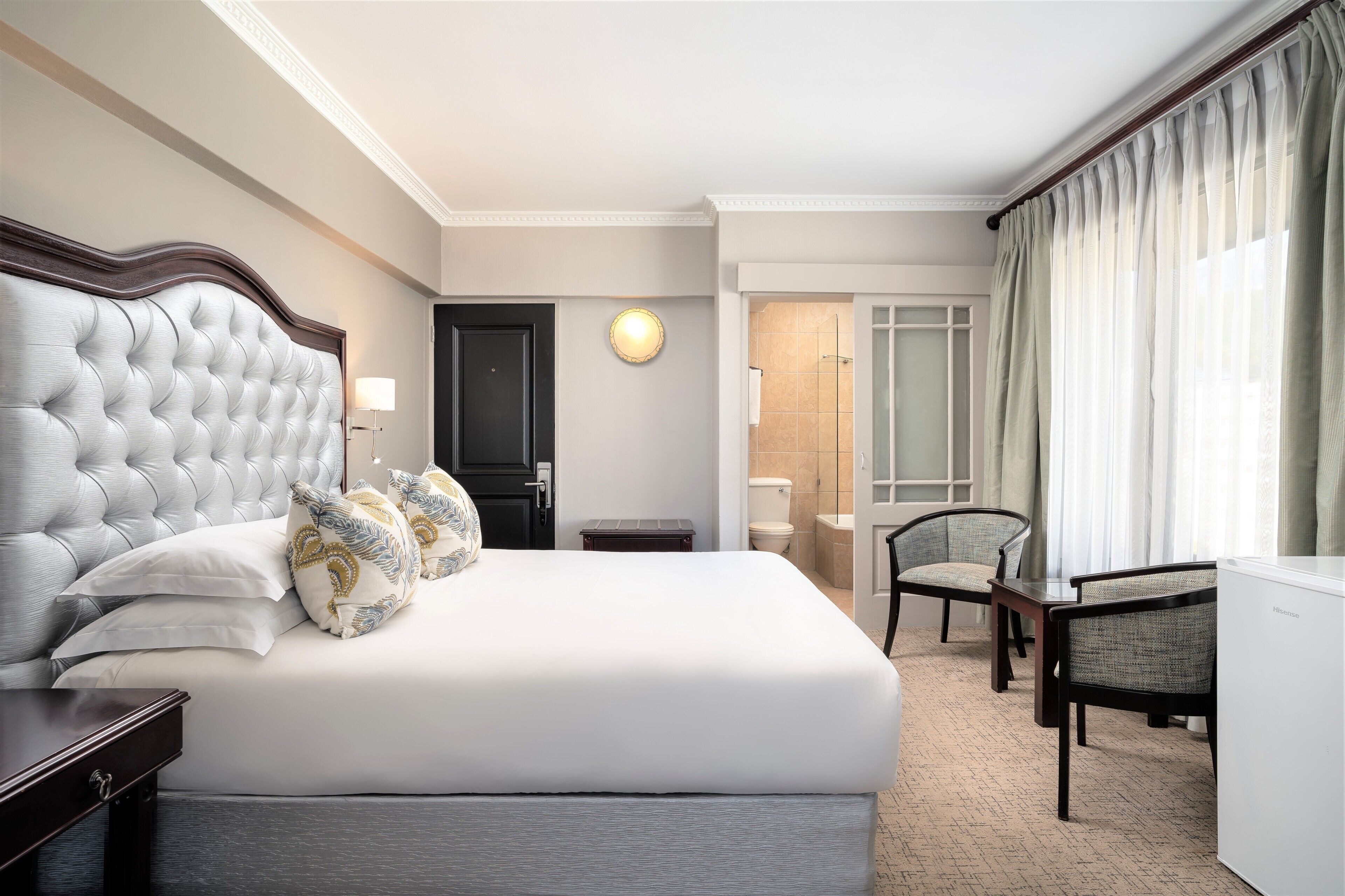 Ein Hotelzimmer mit einem großen Bett, einem Schreibtisch, einem Stuhl, einer Lampe und einem durchgegangenen Tür erkennbaren Badezimmer.