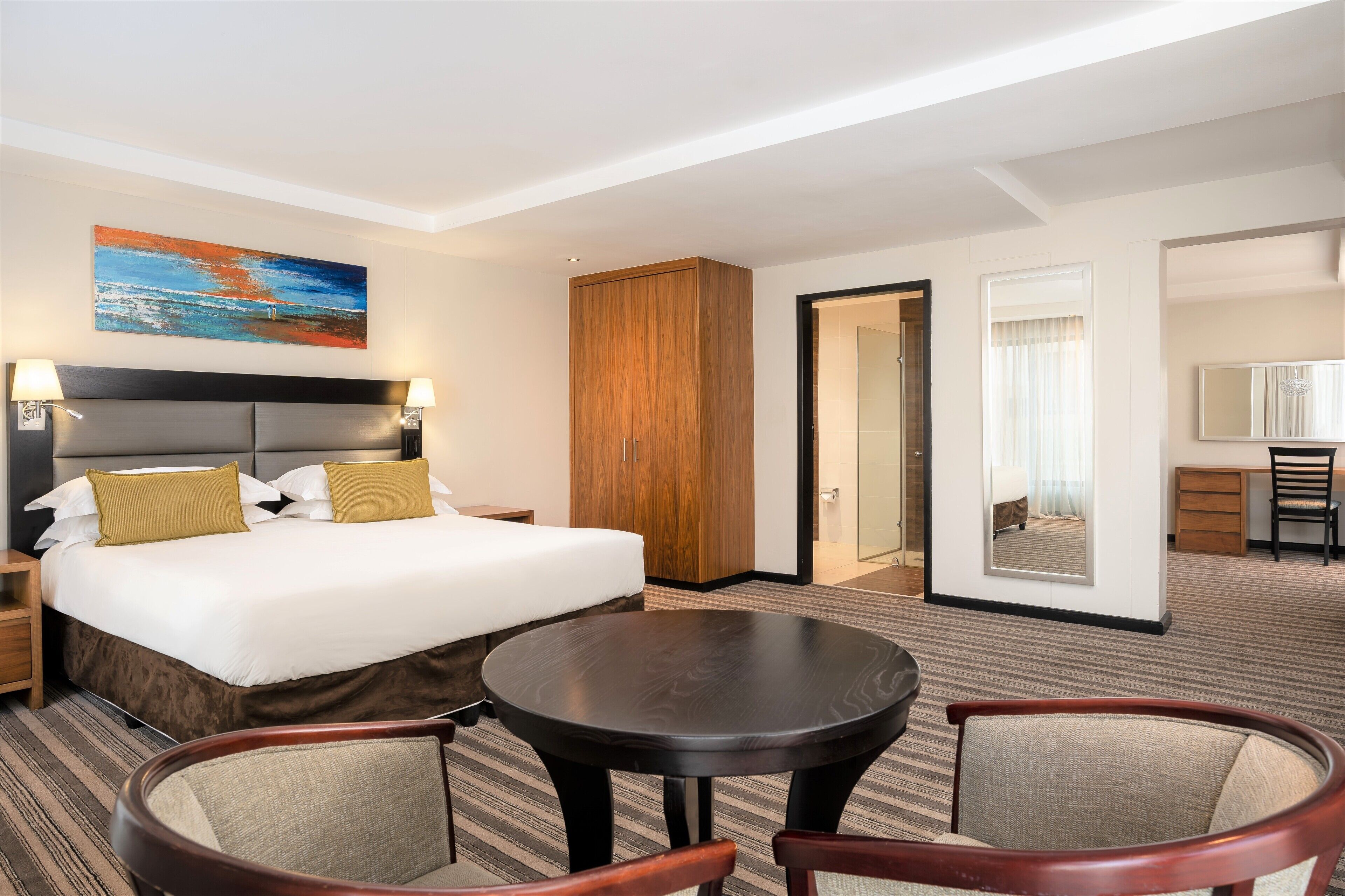A hotel room with a large bed, a wooden wardrobe, a round table, two armchairs, a desk with a chair, and a painting on the wall.