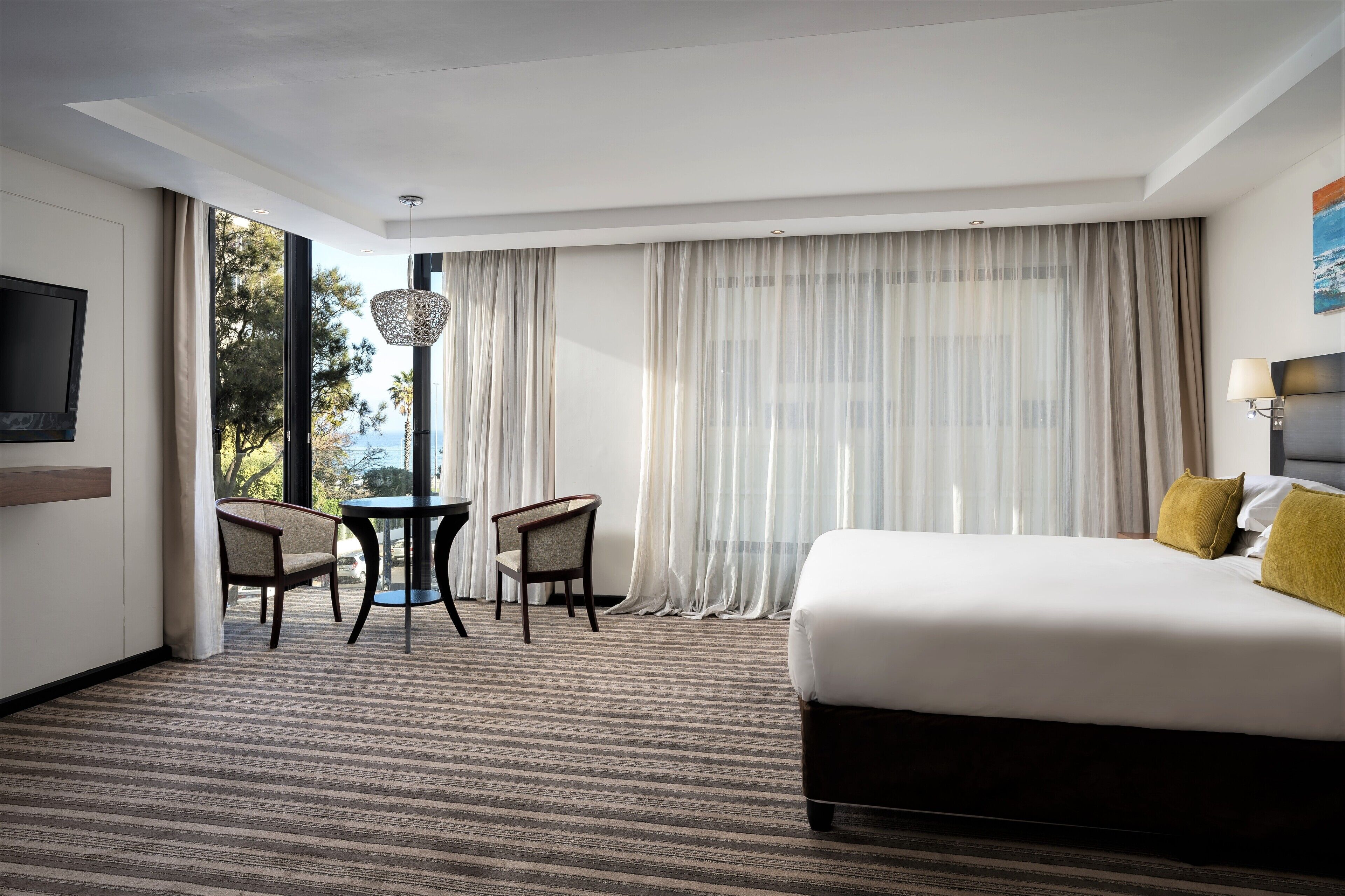 A hotel room with a large bed, a TV on the wall, a dining table with chairs, and a view of trees through sheer curtains.