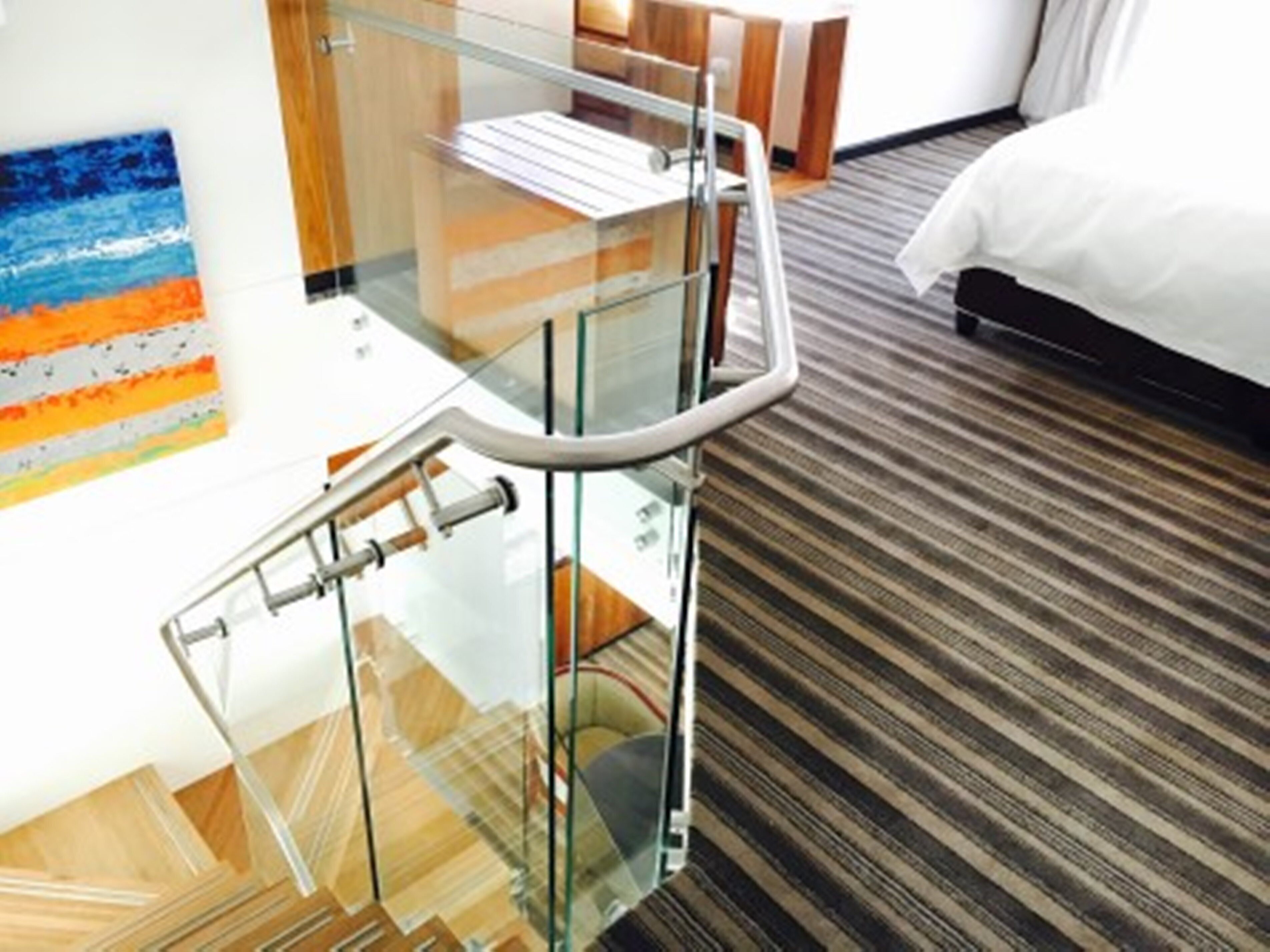 A hotel room with a glass-walled staircase, a bed with white bedding, and a striped carpet.