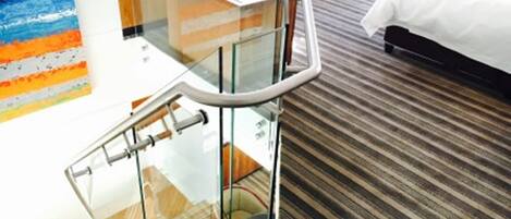 A hotel room with a glass-walled staircase, a bed with white bedding, and a striped carpet.