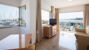 A hotel room with a balcony, a TV, a dining table, and a view of boats and a cityscape.