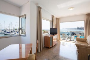 Habitación de hotel con balcón, televisor, mesa de comedor y vistas a barcos y al paisaje urbano.