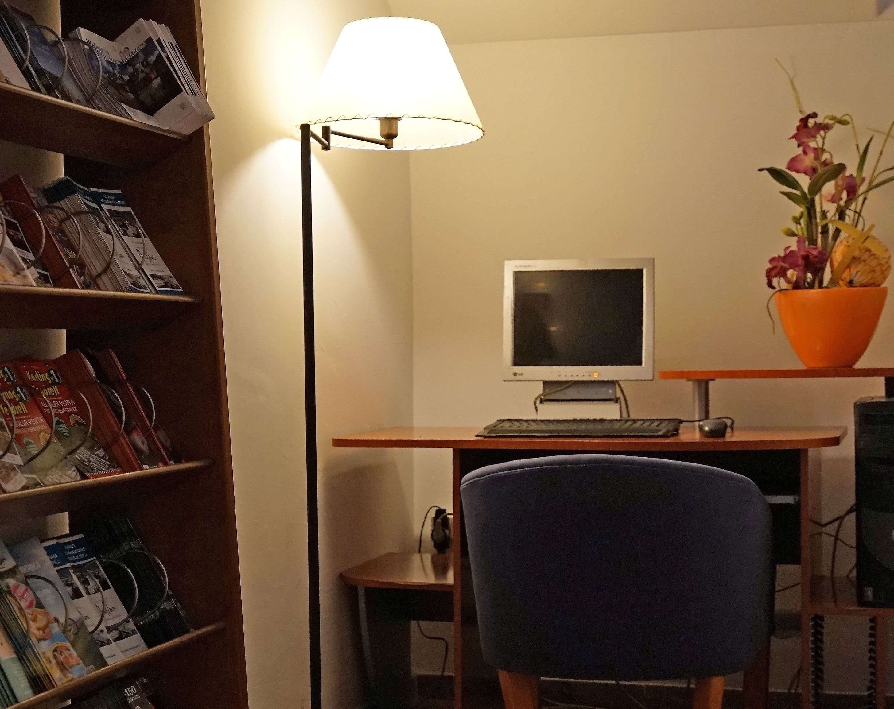 Habitació amb escriptori, monitor d'ordinador, làmpada, cadira i prestatgeria plena de revistes.