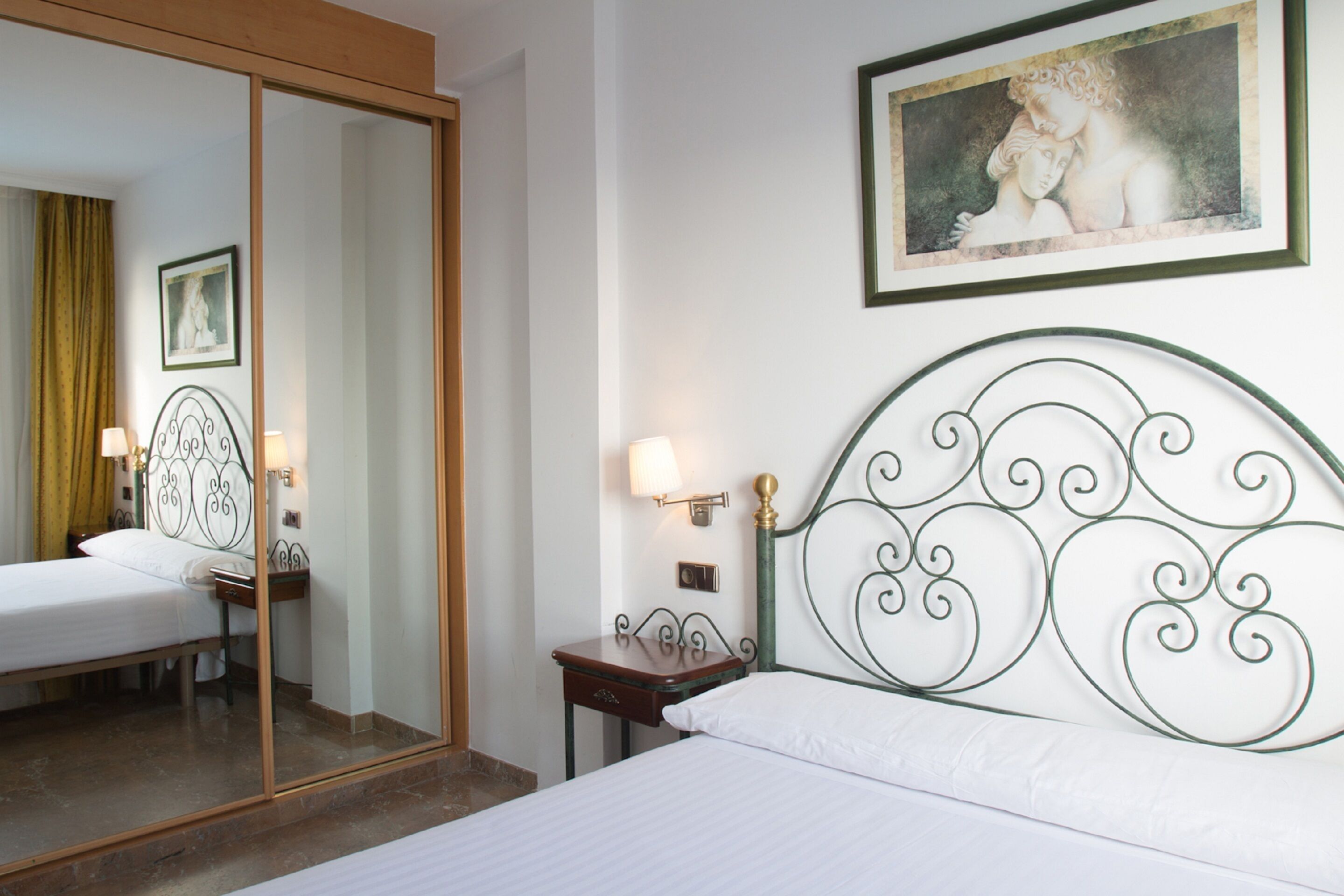 Een hotelkamer met een bed, nachtkastje, spiegel, schilderij en een deur.