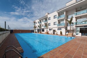 A swimming pool with a tiled deck, surrounded by a multi-story building with balconies and a clear sky.