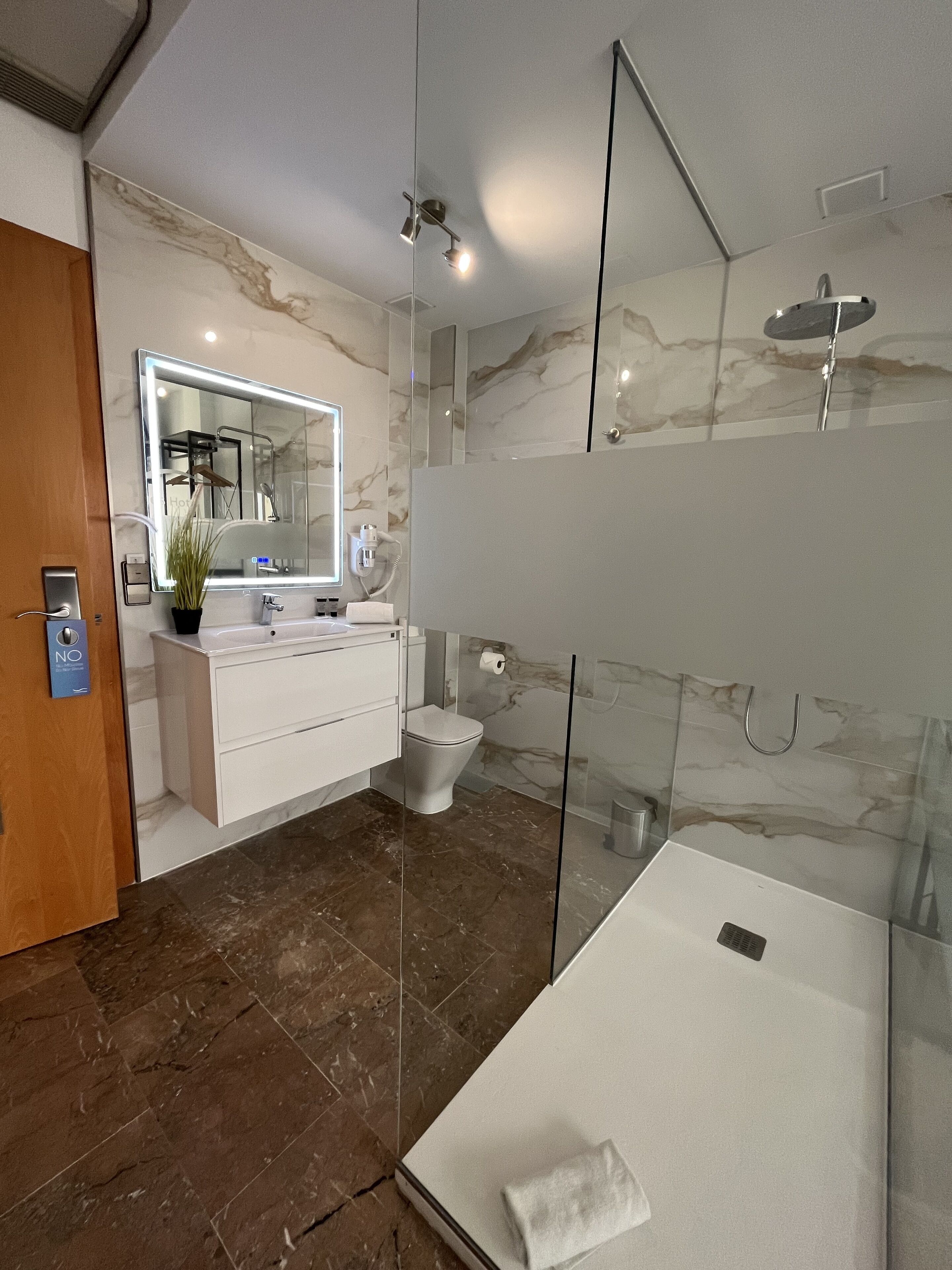 Un baño moderno con ducha envuelta en vidrio, lavamanos blanco y suelo de mármol.