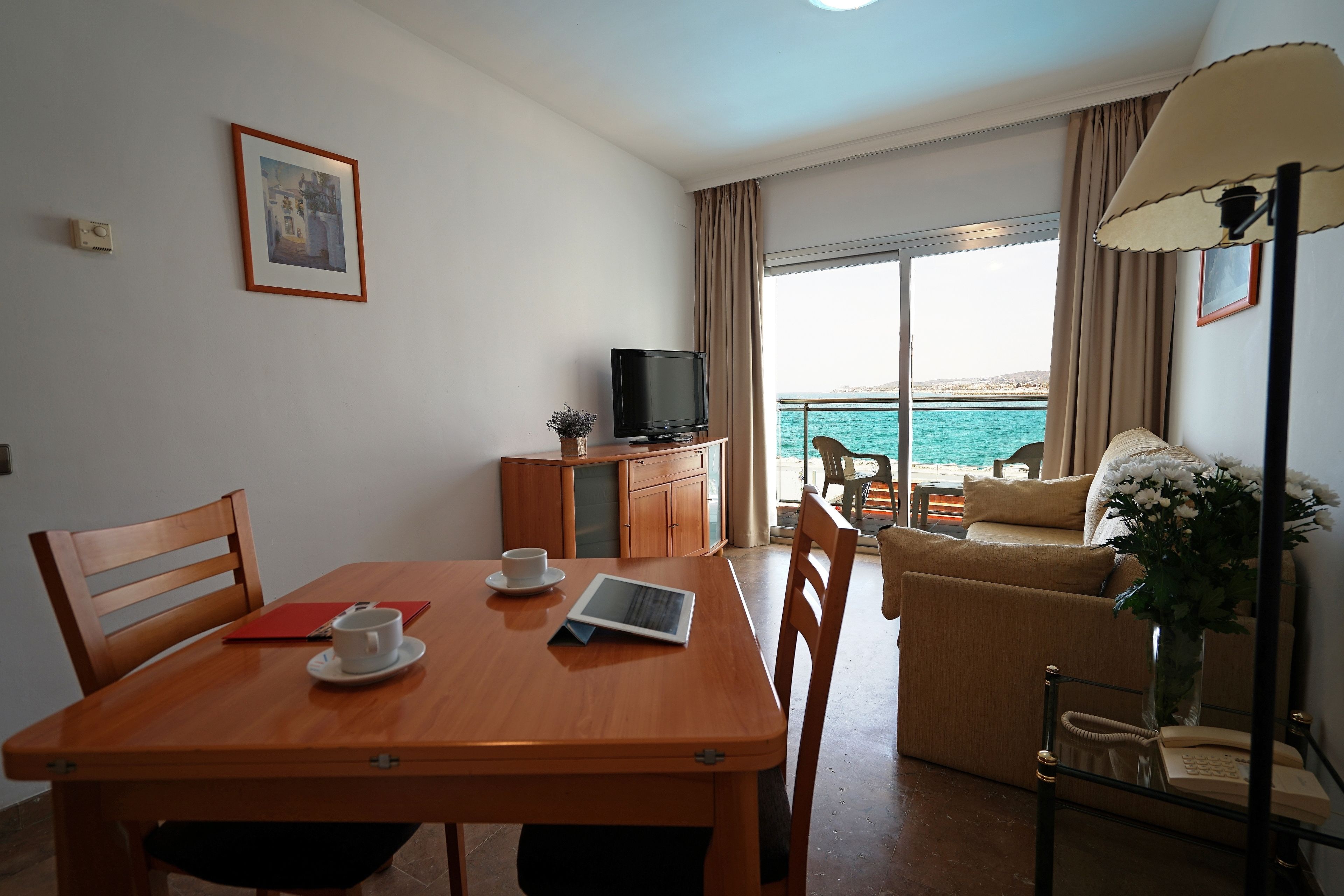 A hotel room with a wooden dining table, chairs, a television, a sofa, and a view of the sea.