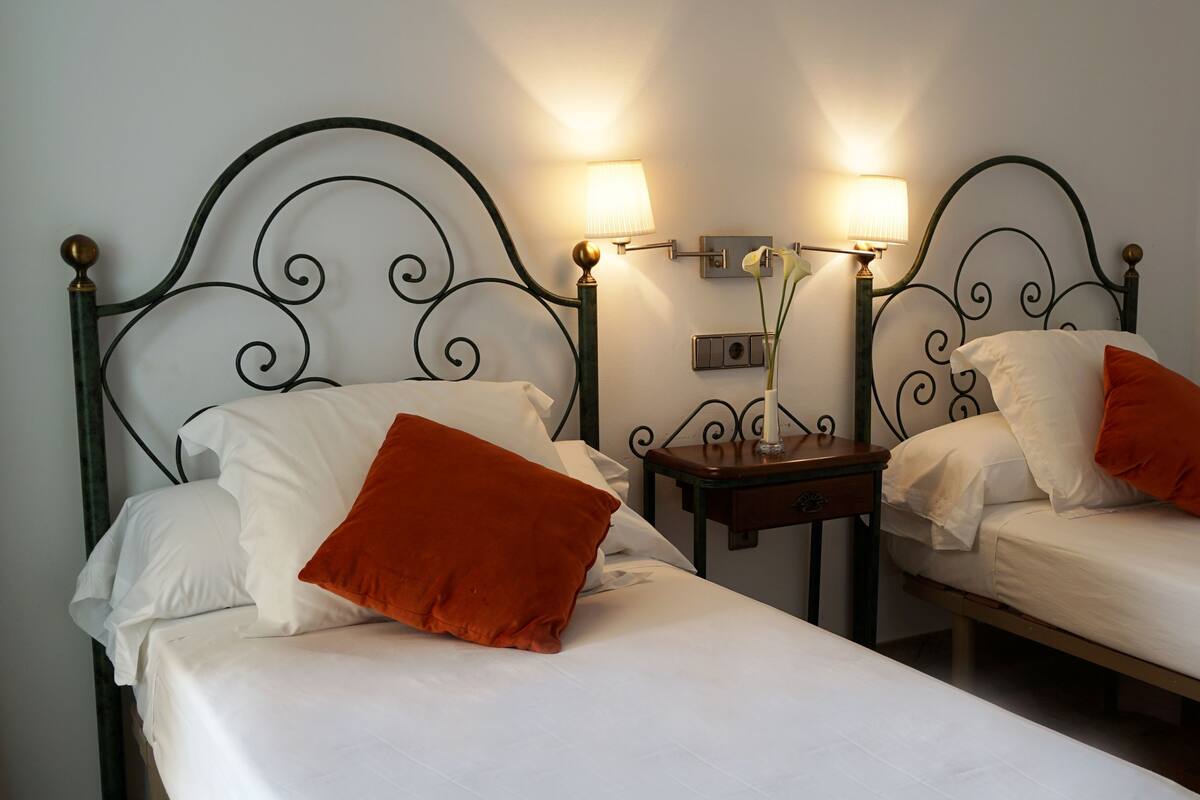 Two single beds with white linens and orange pillows, each with a nightstand and a wall-mounted lamp.