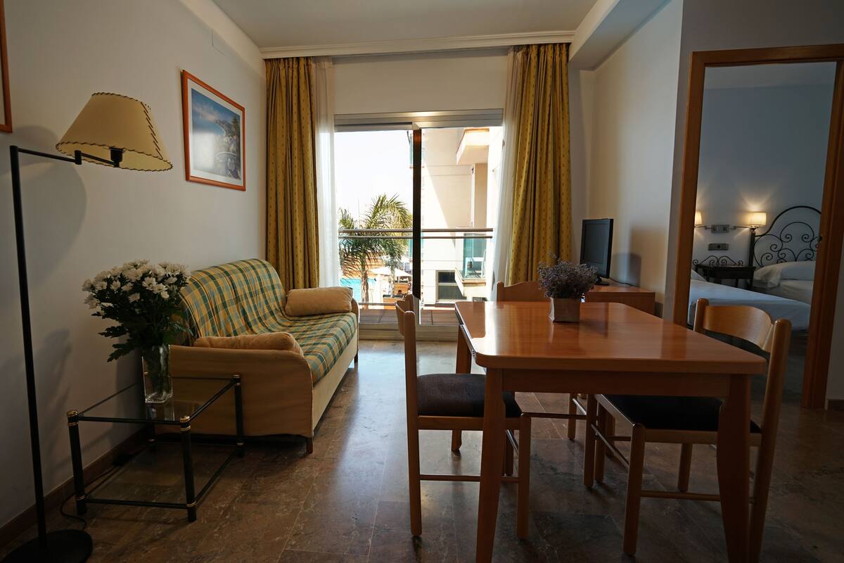 A cozy living room with a sofa, a dining table, and chairs. There is a lamp, a framed picture on the wall, and a window with curtains.