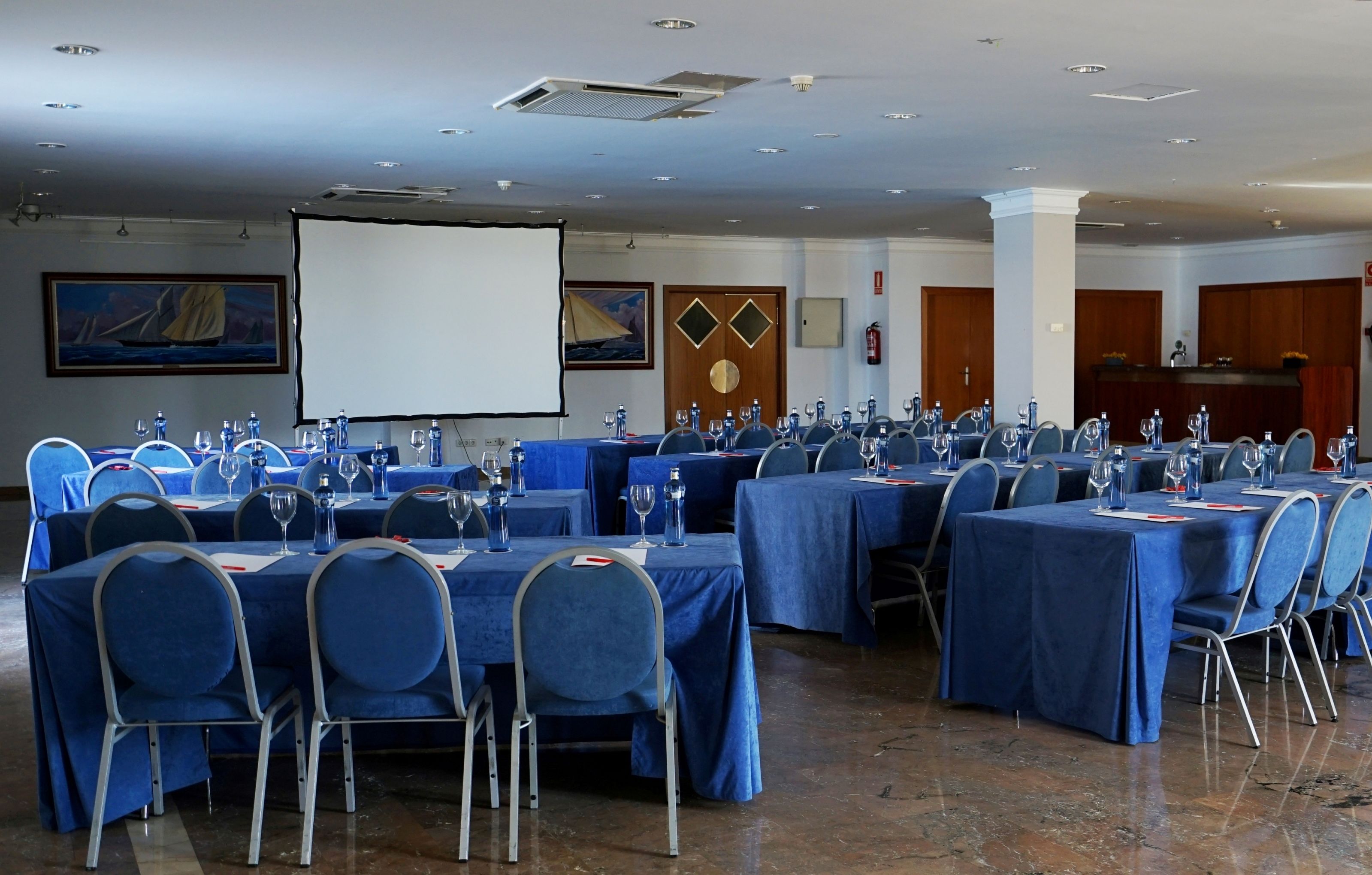 Une salle de conférence avec plusieurs tables disposées pour une réunion, équipée de chaises, de tables et de verres à eau.