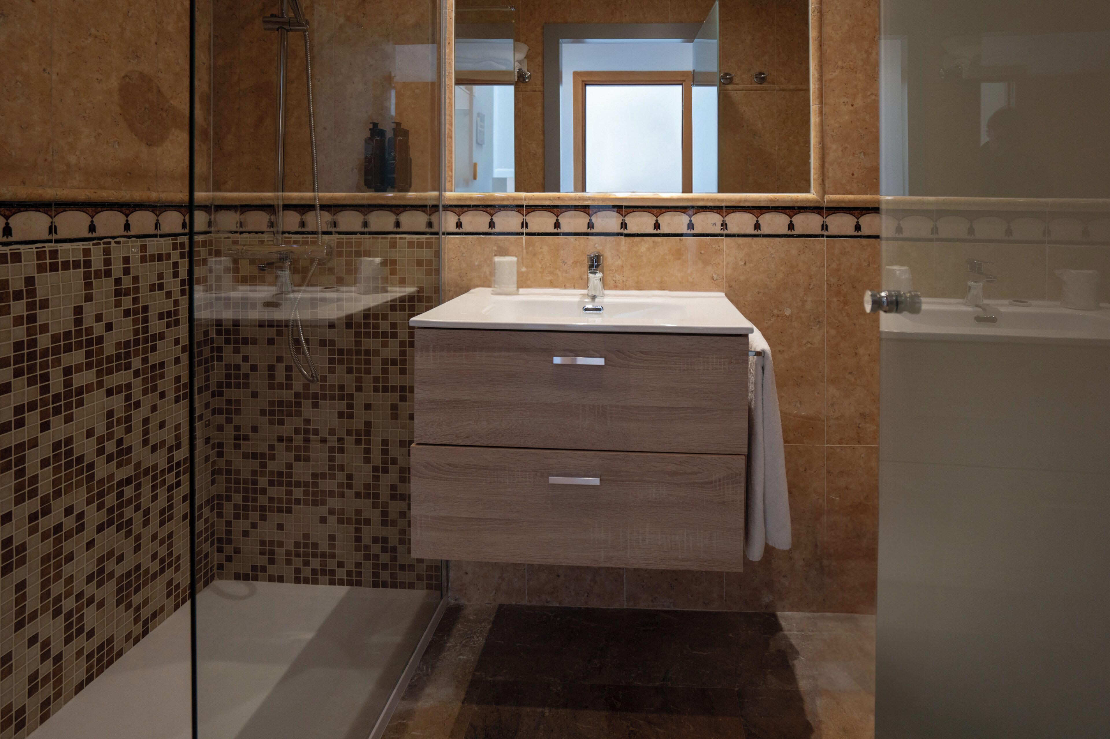 Een badkamer met een douchecabine van glas, een wastafel met spiegel erboven en een betegelde wand met decoratieve rand.
