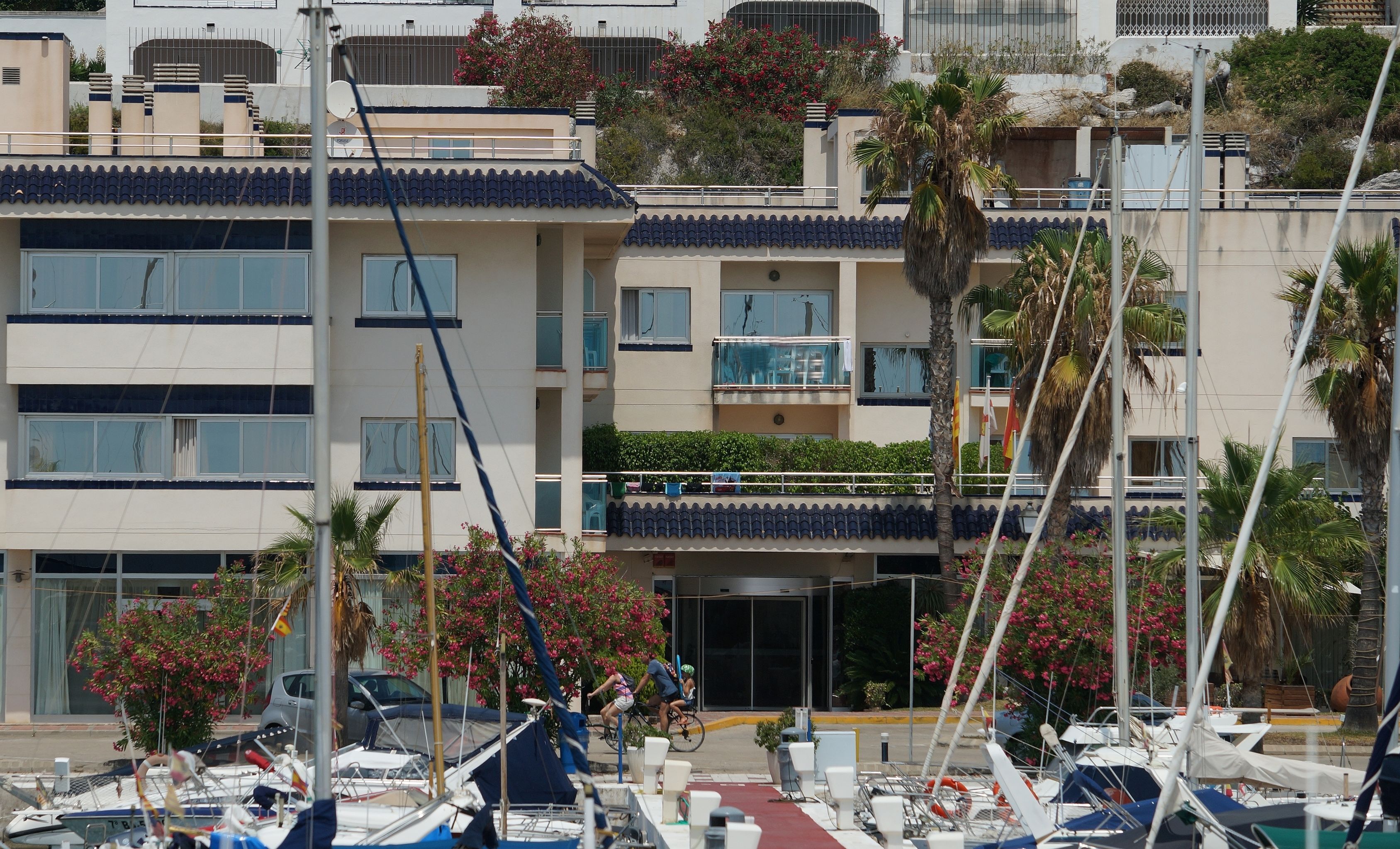 A marina with multiple boats docked, a building with balconies, and a clear sky.