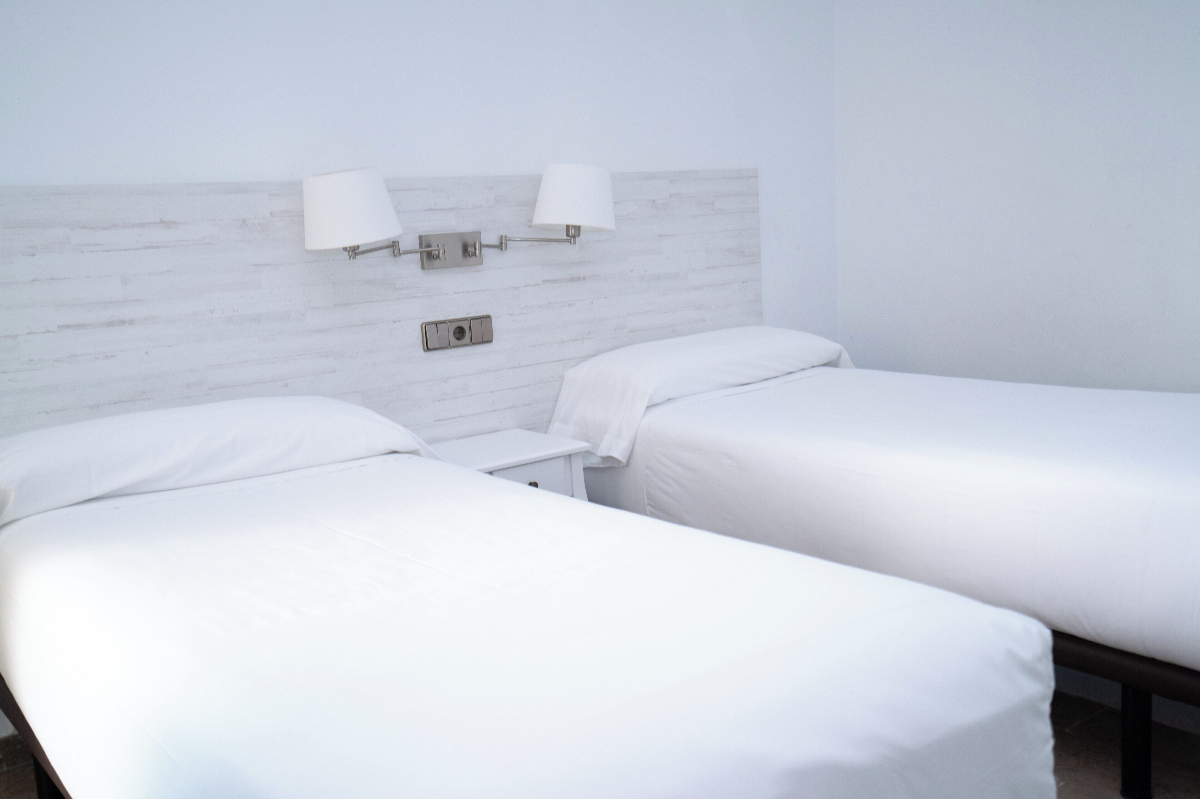 A hotel room with two single beds, a nightstand, a wall-mounted light fixture, and a power outlet.