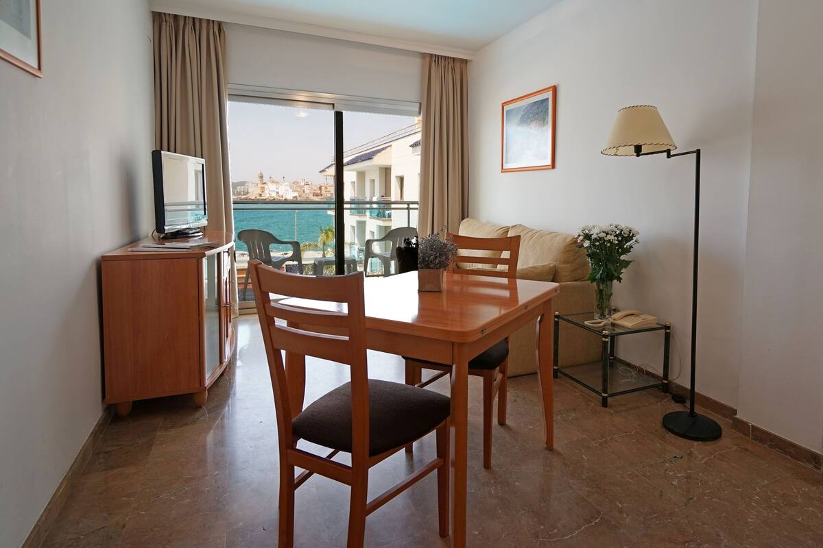 A hotel room with a balcony, a wooden desk with a chair, a sofa, a floor lamp, and a television.