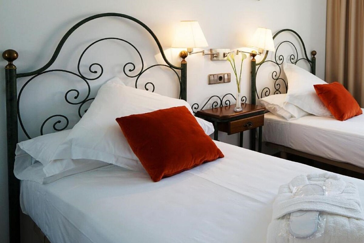 Two beds with white linens, red pillows, and a white robe on the floor.