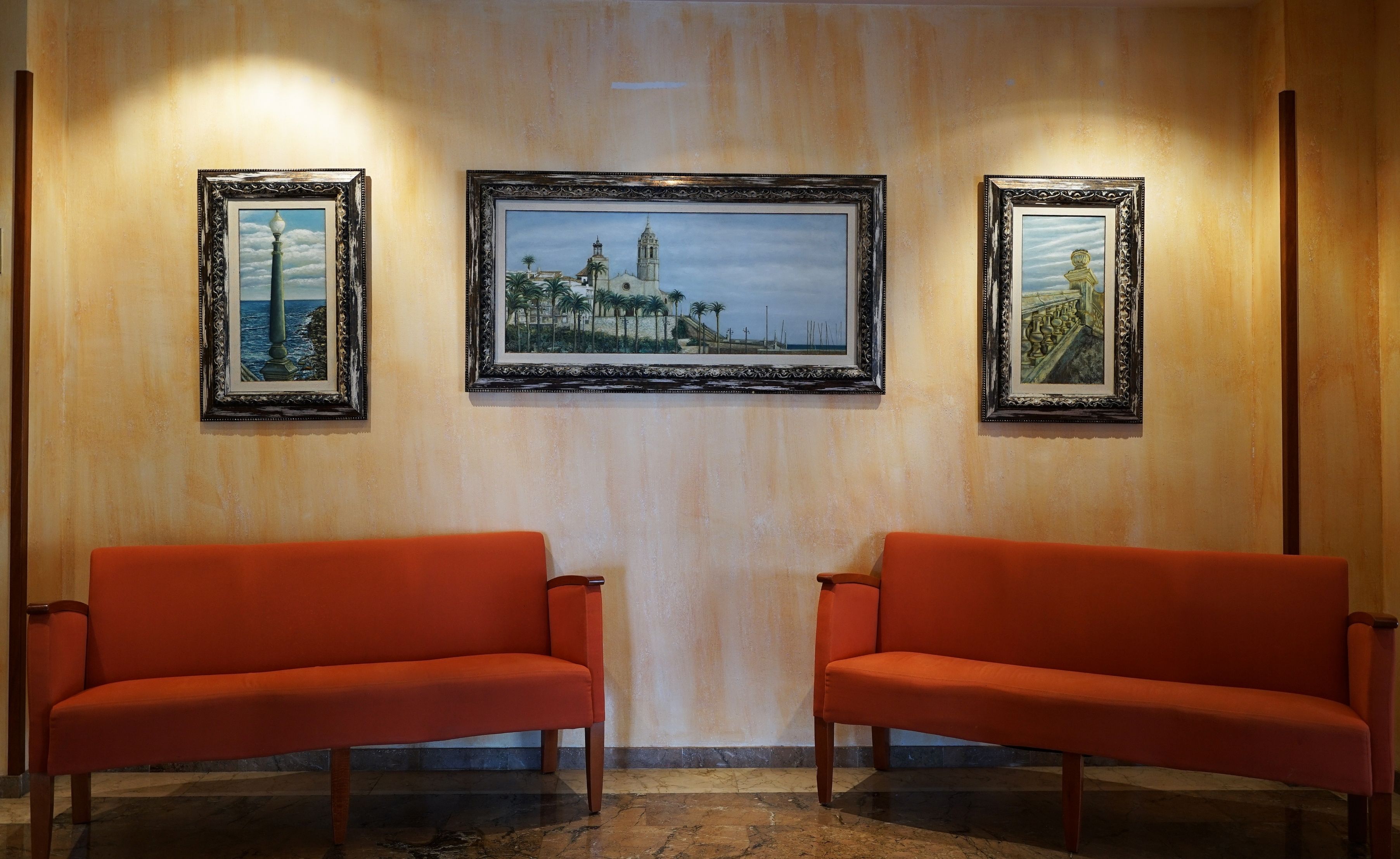 A lobby with two orange sofas, three framed paintings on the wall, and a marble floor.