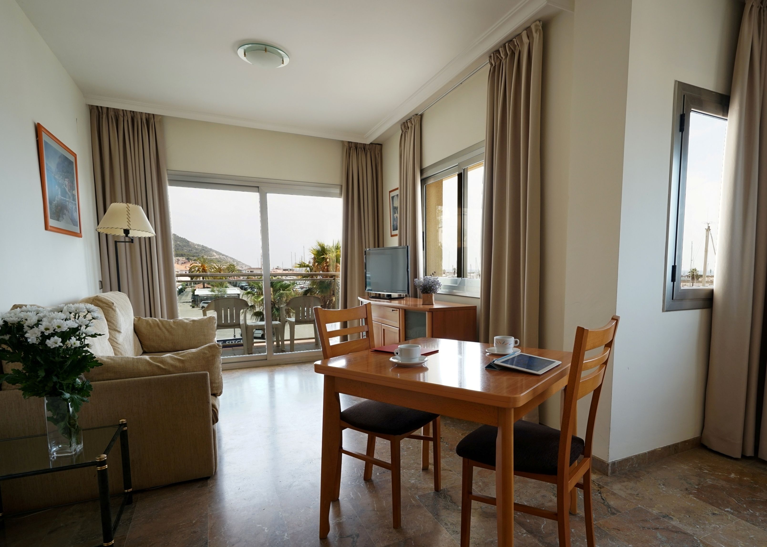 Habitación de hotel con balcón, zona de comedor con mesa y sillas, sofá, televisión y vista panorámica de la ciudad.