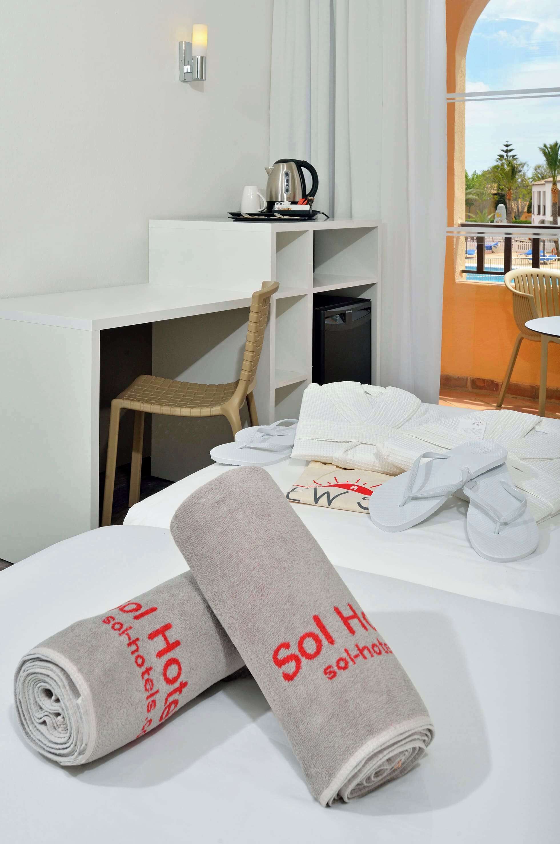 A hotel room with a bed, a desk, a chair, a coffee machine, and towels with "Sol Hotel" branding.