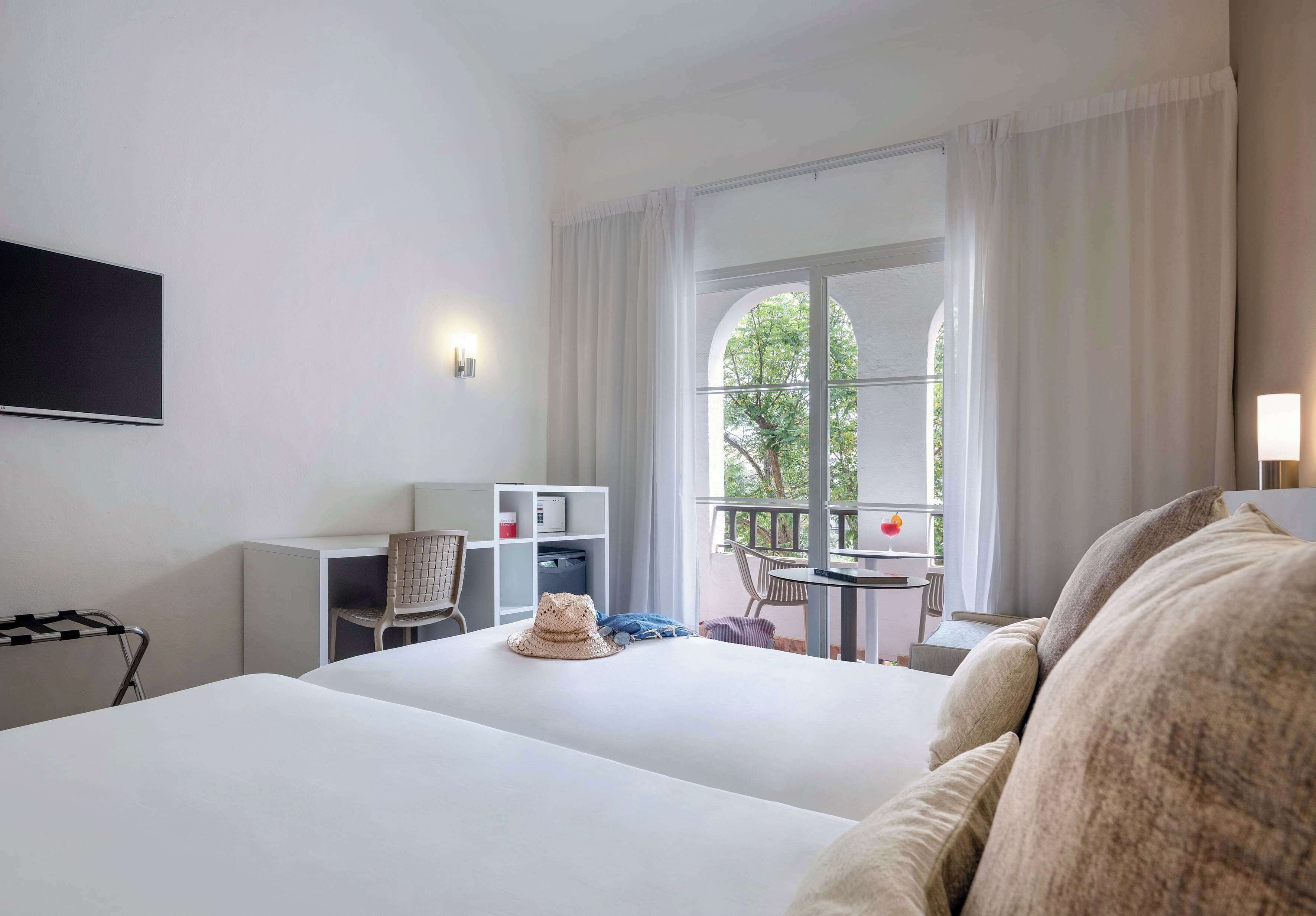 A hotel room with two beds, a desk, a chair, a small table, and a balcony with a view of trees.