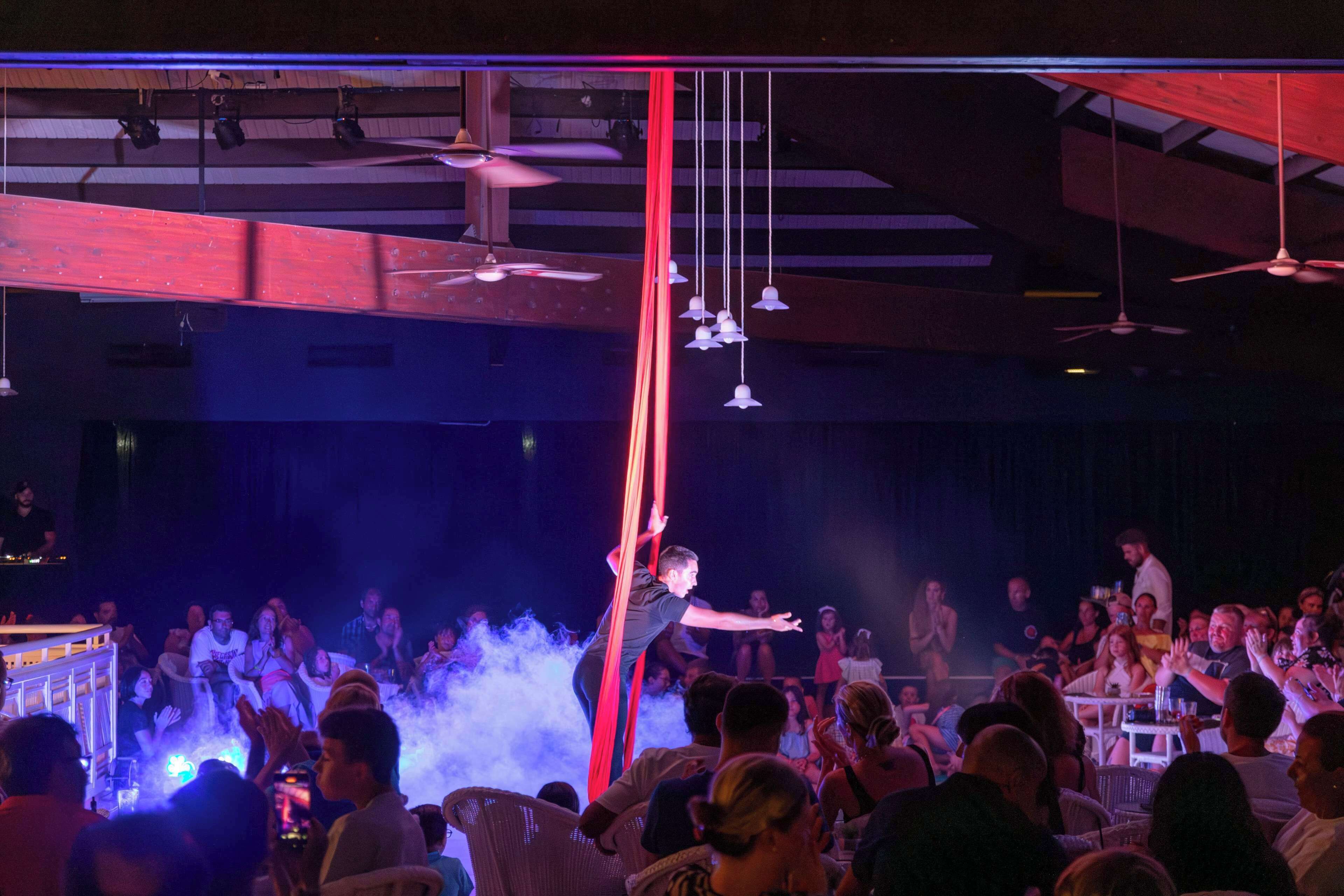 A performance with an audience seated at tables, a stage with a performer, and a ceiling with hanging lights and fans.