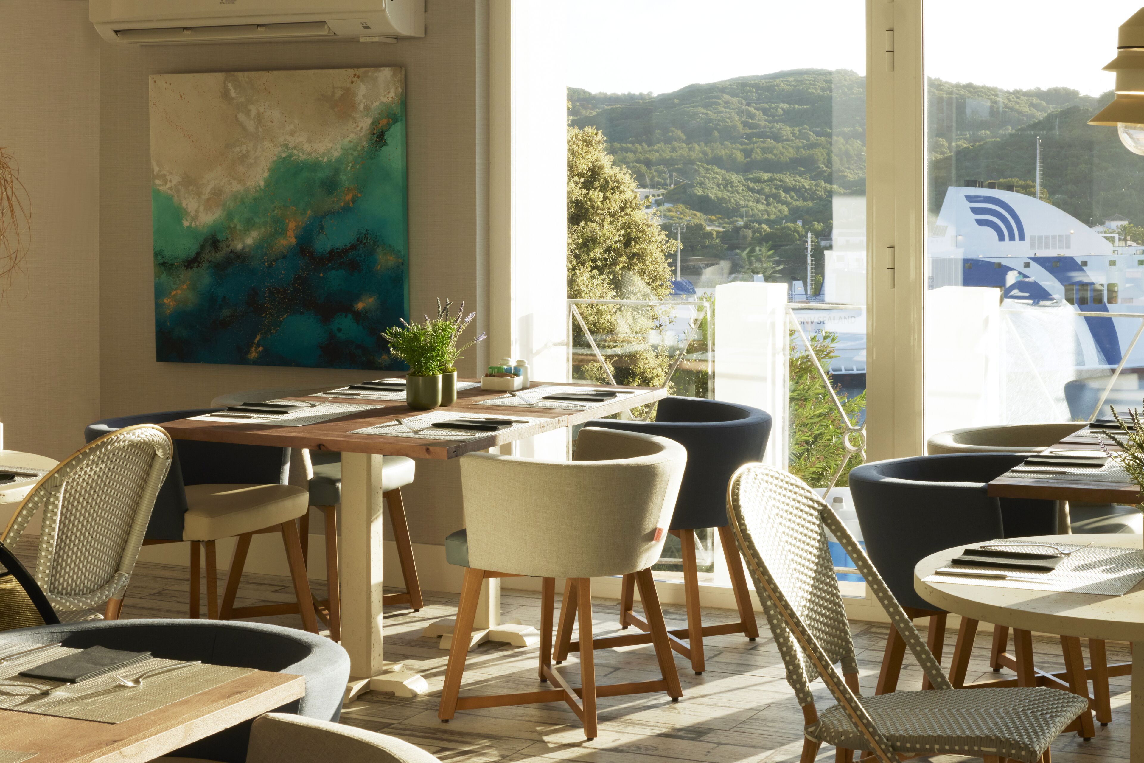 A dining area with tables and chairs, a large abstract painting on the wall, and a view of a boat and hills outside.