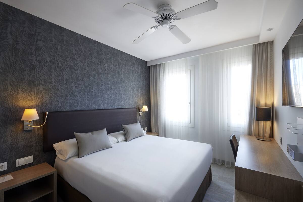 A modern hotel room with a large bed, a desk, a ceiling fan, and a window with curtains.