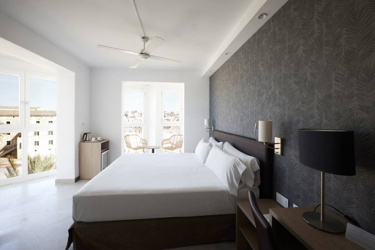 A modern hotel room with a large bed, a ceiling fan, a desk with a lamp, and a view of the city through large windows.