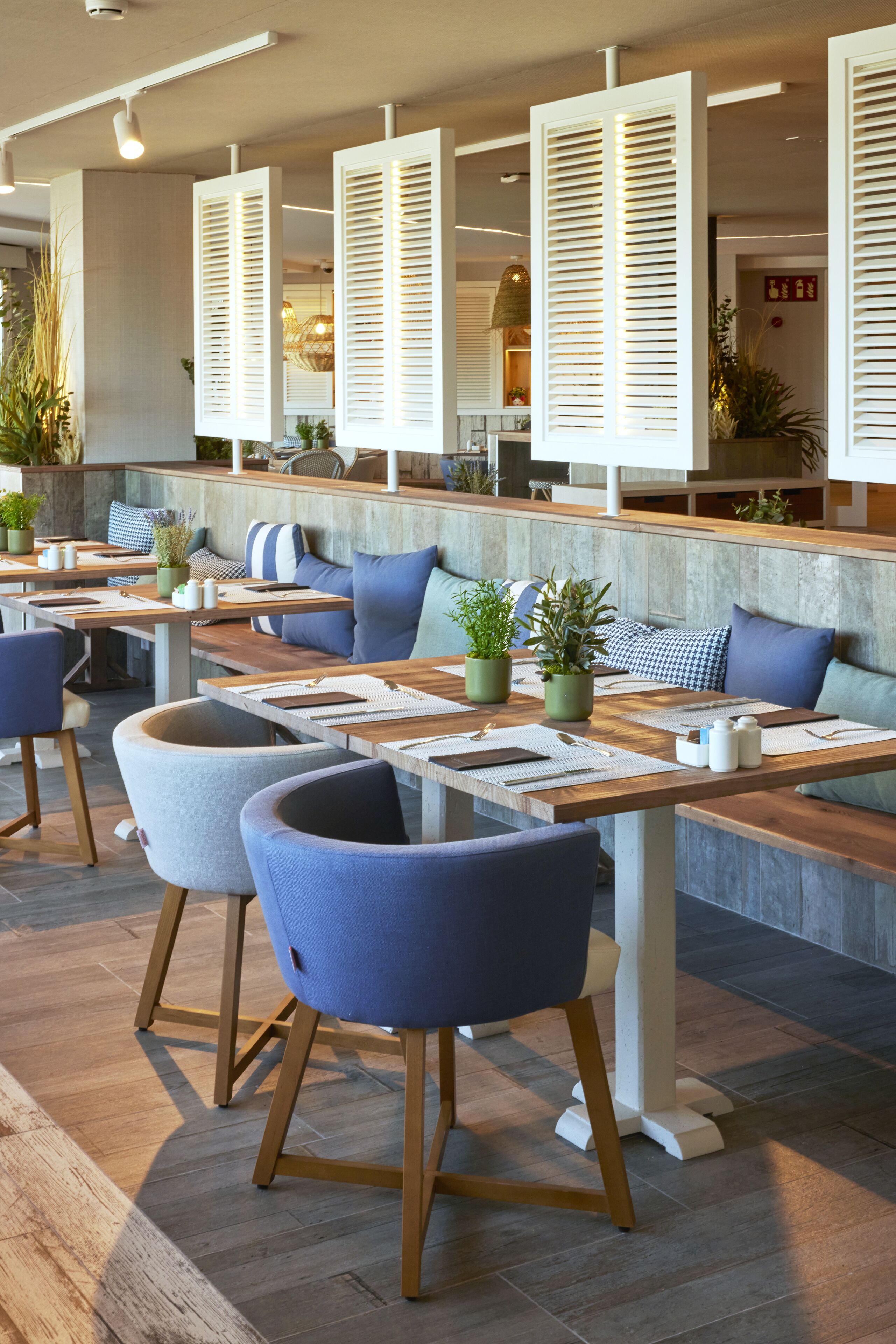 A modern restaurant with wooden tables and blue chairs, featuring a seating area with cushions and potted plants.