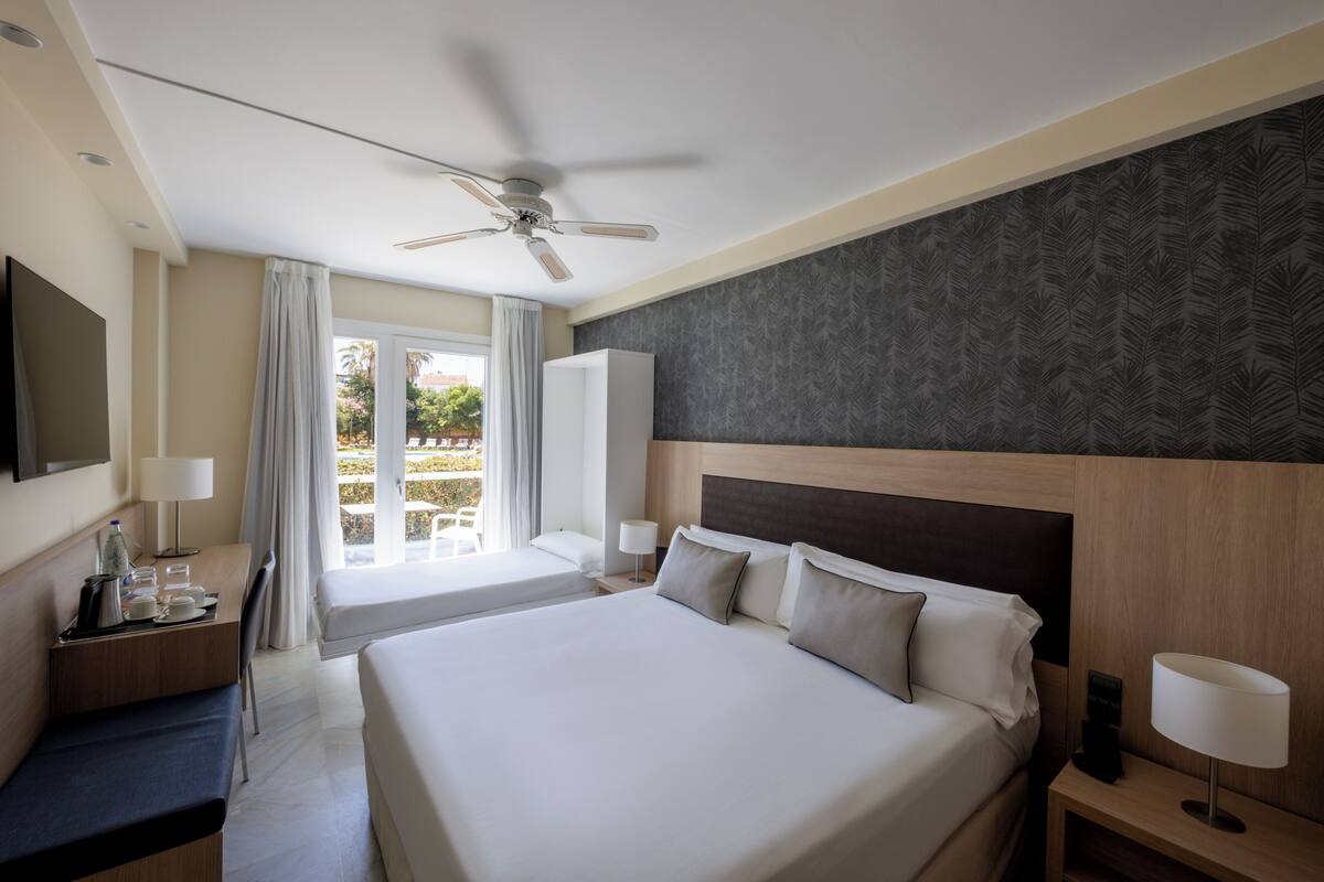 A hotel room with a large bed, a desk with a chair, a TV, a ceiling fan, and a window with curtains.