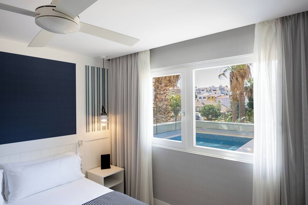 A hotel room with a bed, a nightstand, a ceiling fan, a window with a view of a swimming pool and buildings.